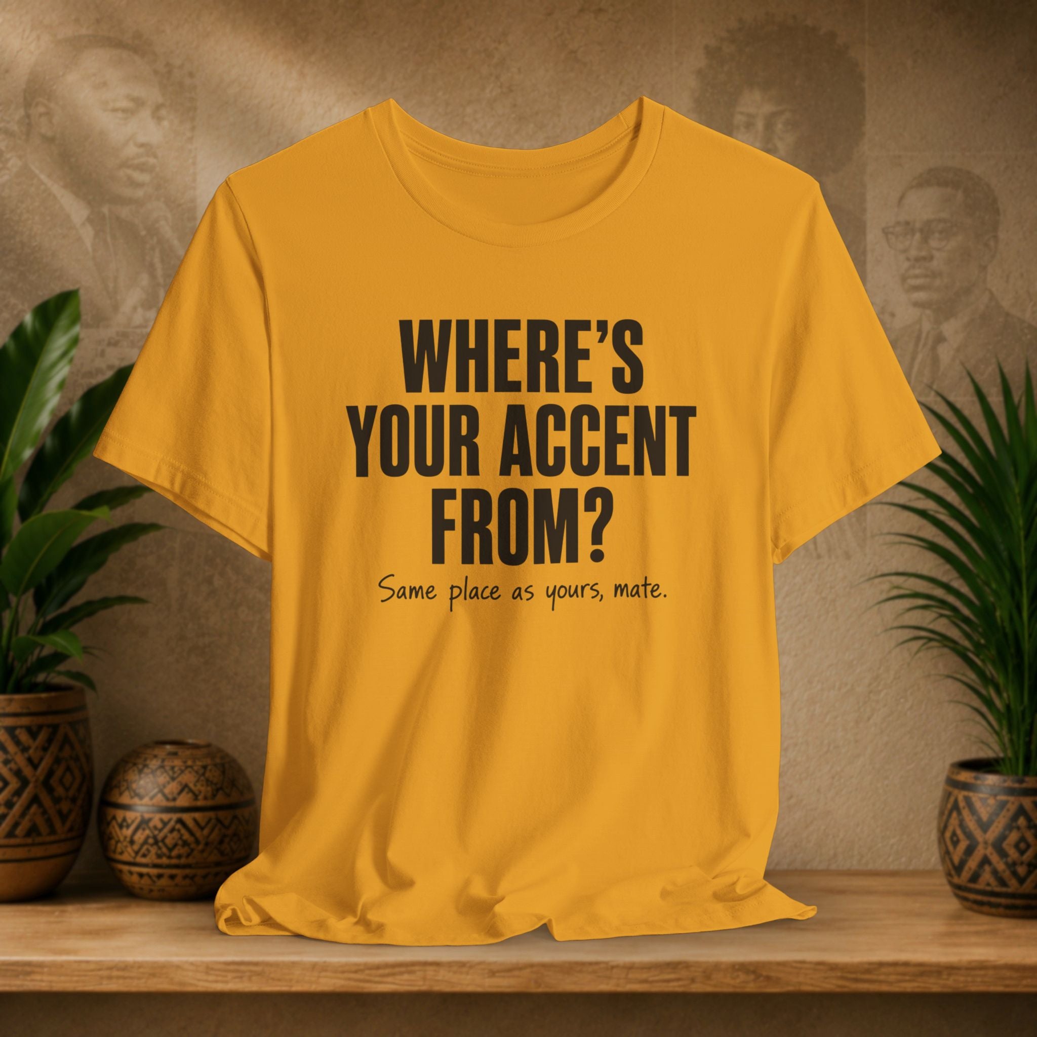 Yellow t-shirt with text 'WHERE'S YOUR ACCENT FROM? Same place as yours, mate.' on a wooden surface with plants and a textured wall background.
