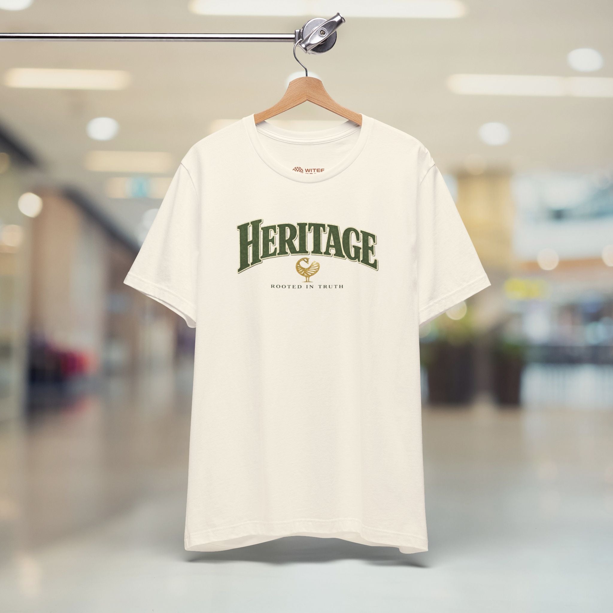 Beige t-shirt with 'HERITAGE' text on a hanger in a blurred indoor setting