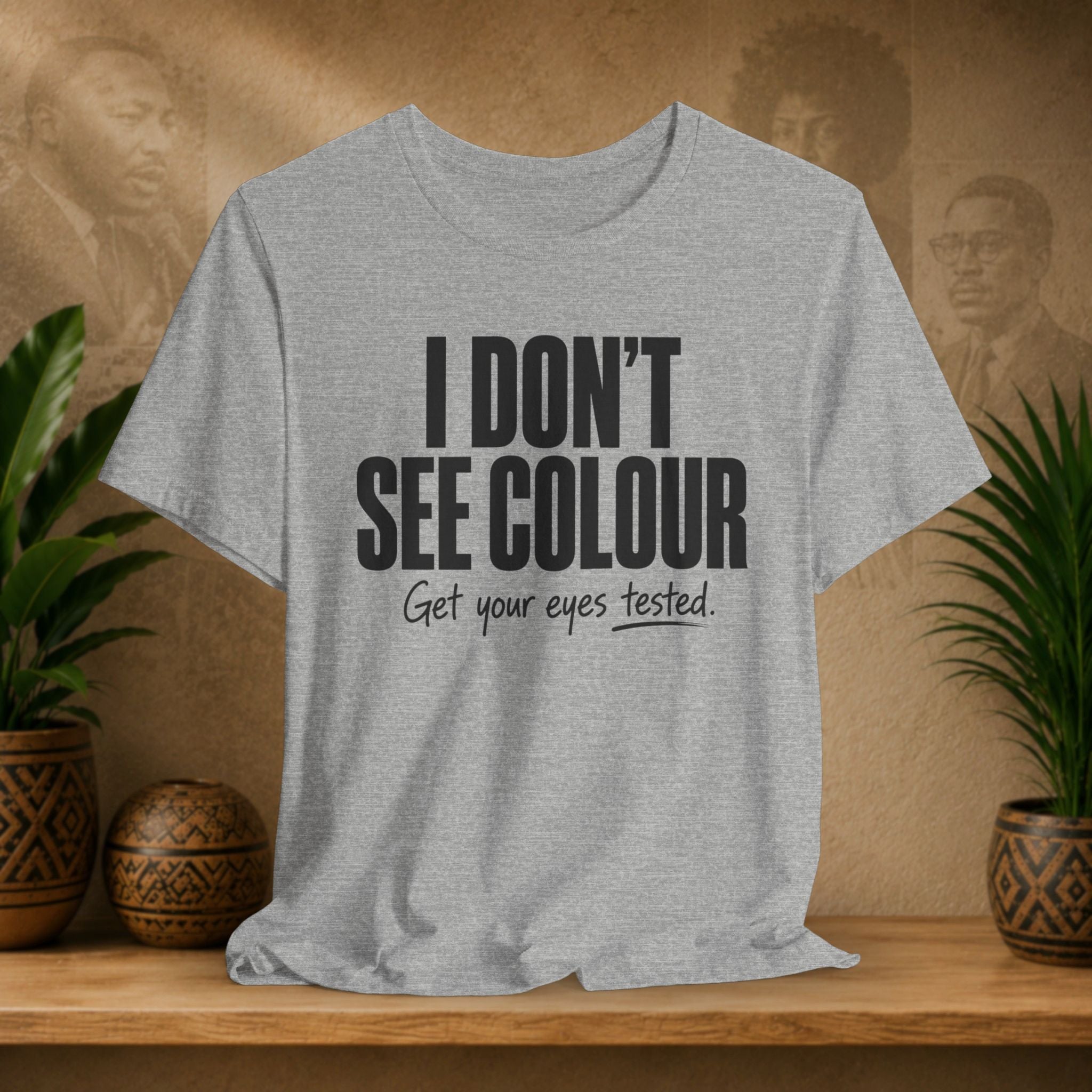Gray t-shirt with text 'I DON'T SEE COLOUR Get your eyes tested.' on a wooden surface with plants and a blurred background.