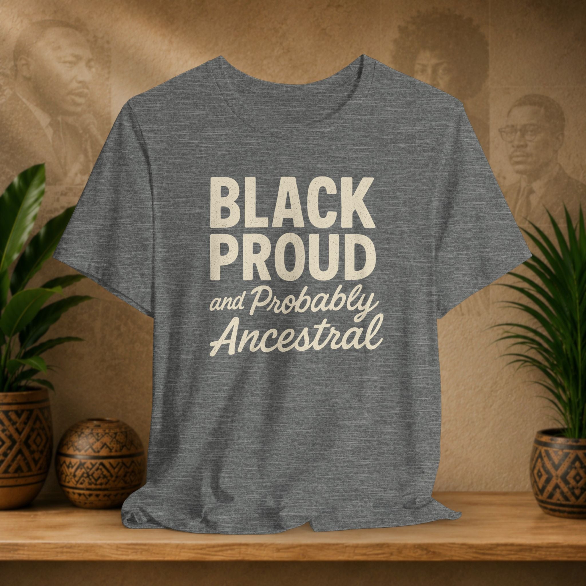 Gray t-shirt with 'Black Proud and Probably Ancestral' text on a wooden surface with plants and decorative items.