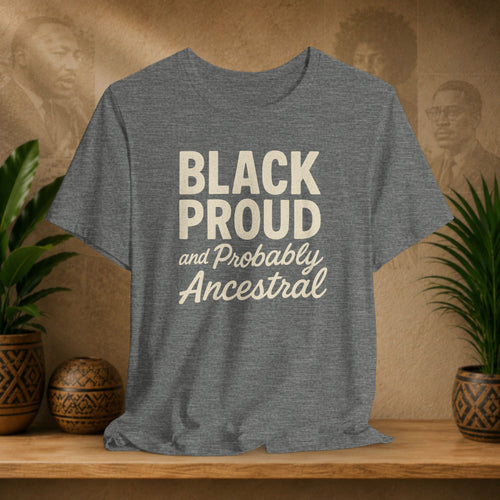Gray t-shirt with 'Black Proud and Probably Ancestral' text on a wooden surface with plants and decorative items.