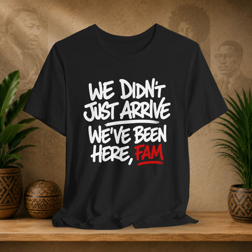 Black t-shirt with white and red text on a wooden surface with plants and a textured wall in the background