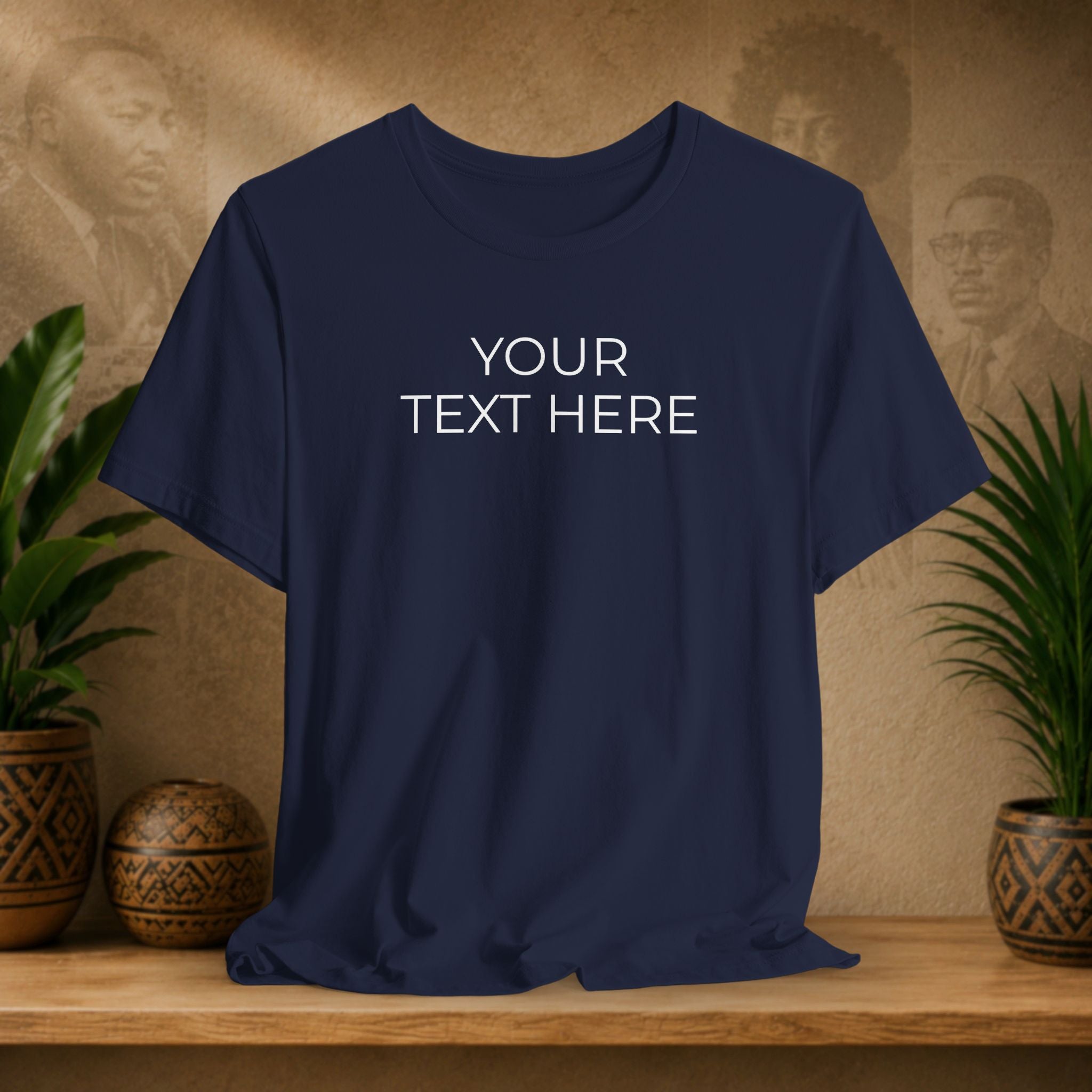 A navy blue short-sleeve t-shirt with 'YOUR TEXT HERE' printed in white on the chest, displayed on a wooden shelf with decorative plants and textured background.