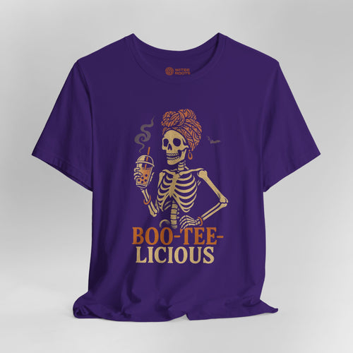 A purple short-sleeve t-shirt with a graphic of a skeleton holding a beer mug, with the text 'BOO-TEE-LICIOUS' printed below in orange and beige. The shirt is displayed against a neutral gray background.
