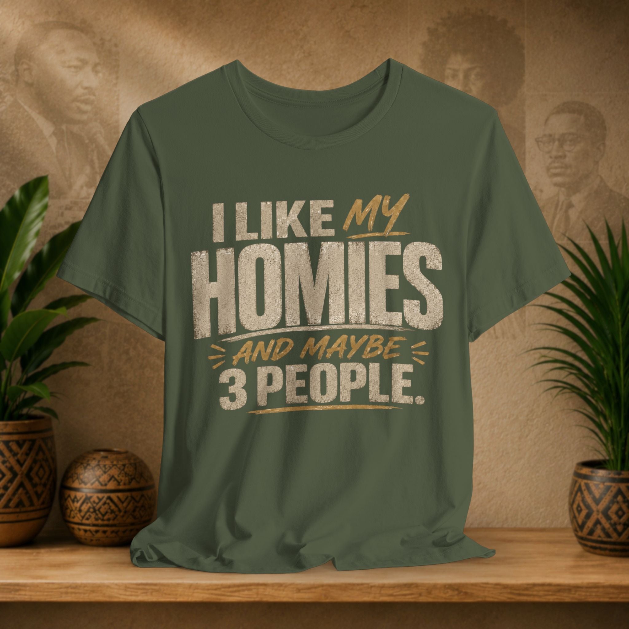 A military green graphic t-shirt displayed on a wooden shelf. The shirt features a humorous slogan printed in white and tan lettering: 'I LIKE MY HOMIES and maybe 3 PEOPLE.