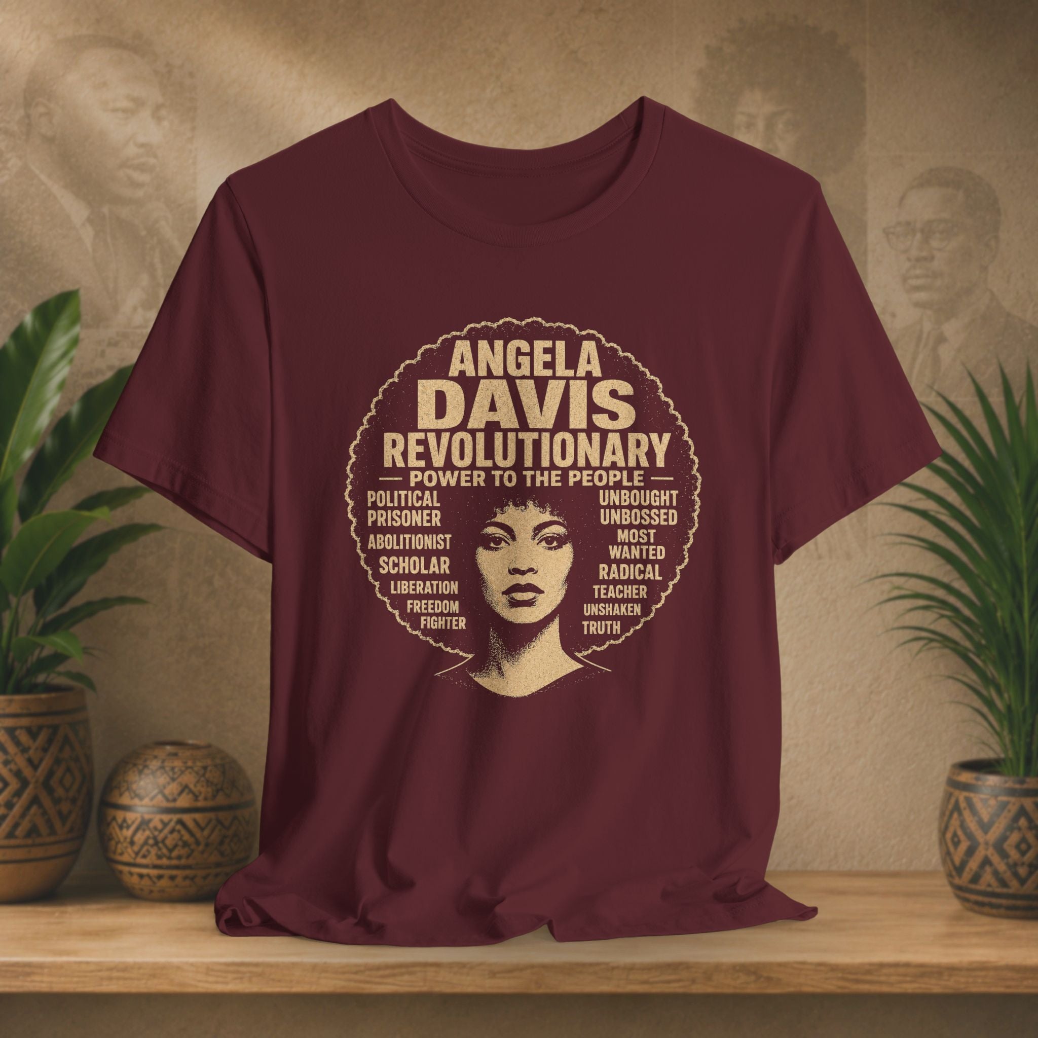 A maroon short-sleeve graphic T-shirt displayed on a wooden surface with potted plants and a textured wall behind it. The shirt features a large circular graphic in tan and cream with the text 'ANGELA DAVIS REVOLUTIONARY' and a stylized portrait of Angela Davis.