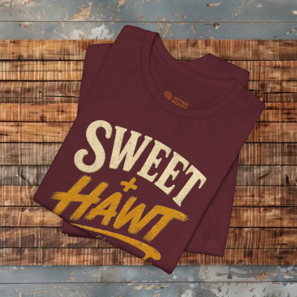 A folded maroon t-shirt with a graphic reading 'SWEET + HAWT' in cream and mustard yellow, displayed on a rustic wooden plank surface.
