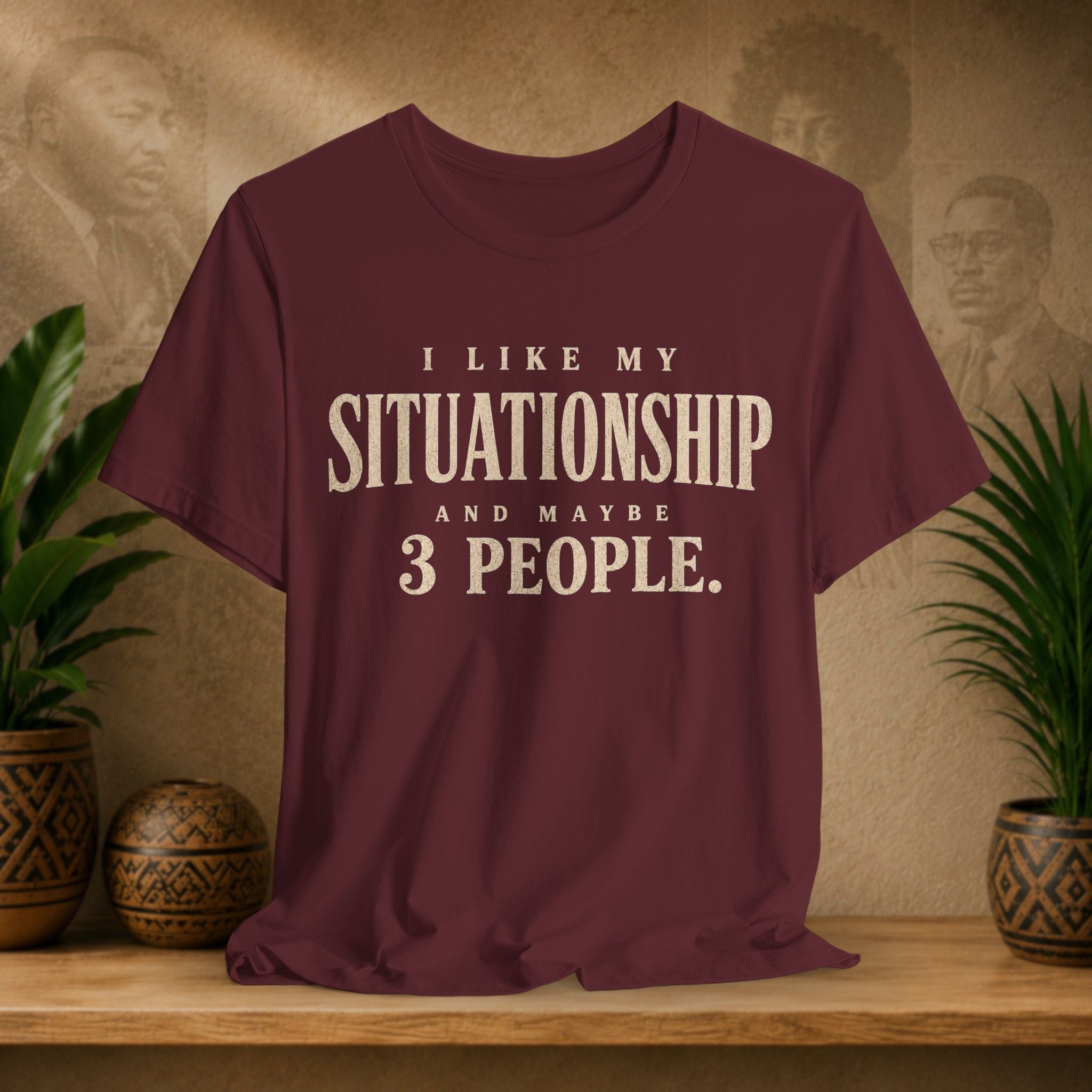 A maroon short-sleeve graphic t-shirt with cream text reading 'I LIKE MY SITUATIONSHIP AND MAYBE 3 PEOPLE.' displayed on a wooden shelf with potted plants and a textured background.