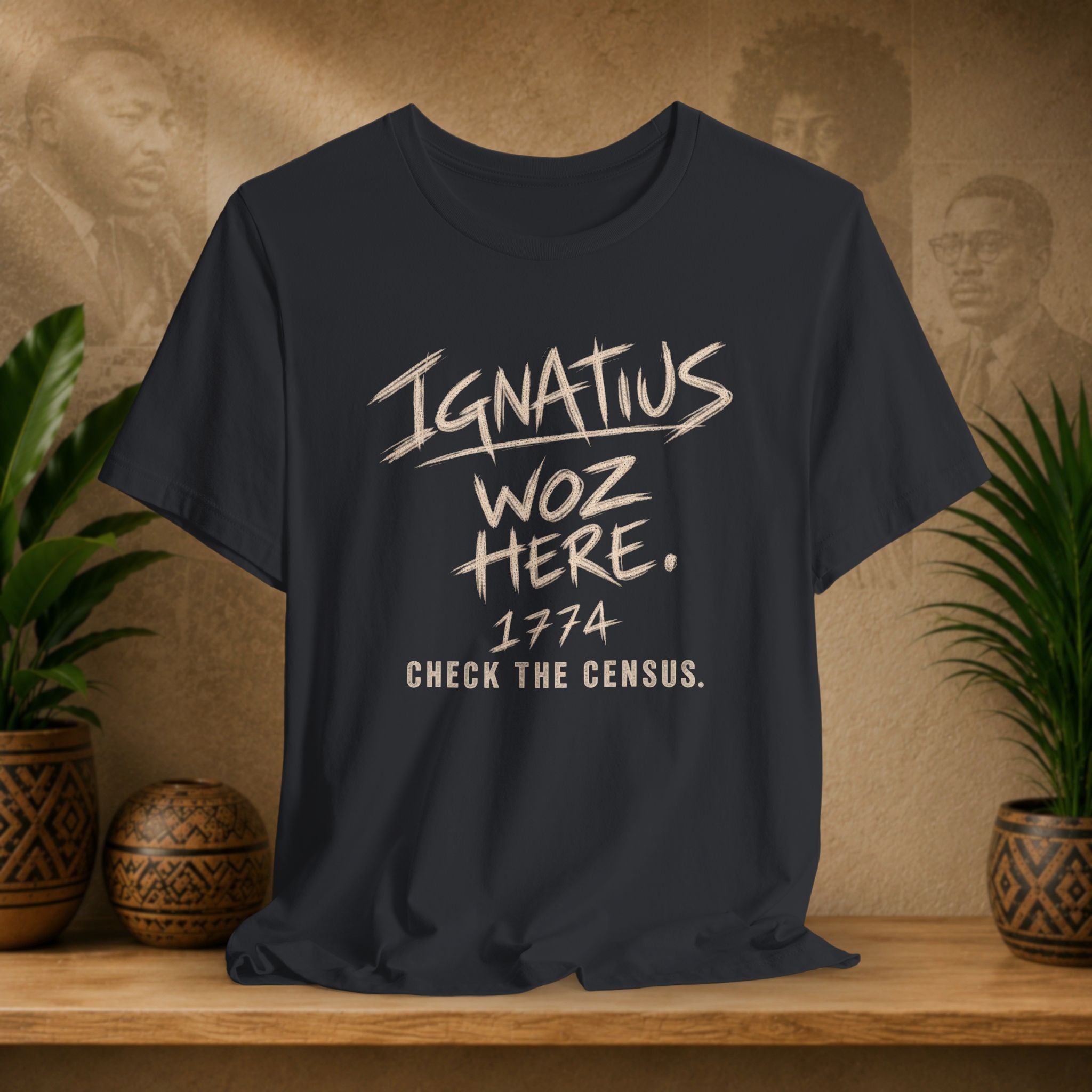 A black graphic t-shirt displayed on a wooden shelf, flanked by potted plants and a textured background with sepia portraits. The shirt features a distressed cream-colored text graphic reading 'Ignatius Woz Here.