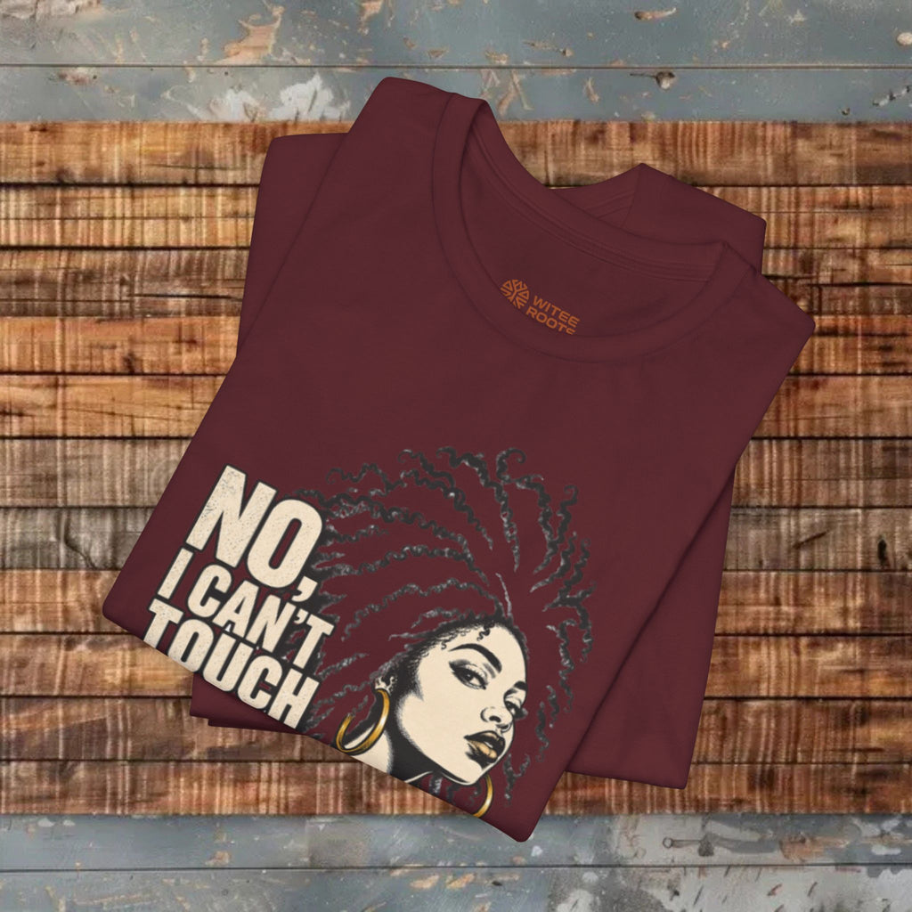 A folded maroon t-shirt with a graphic print of a woman with an afro and the text 'NO, I CAN'T TOUCH' in beige, displayed on a rustic wooden plank background.