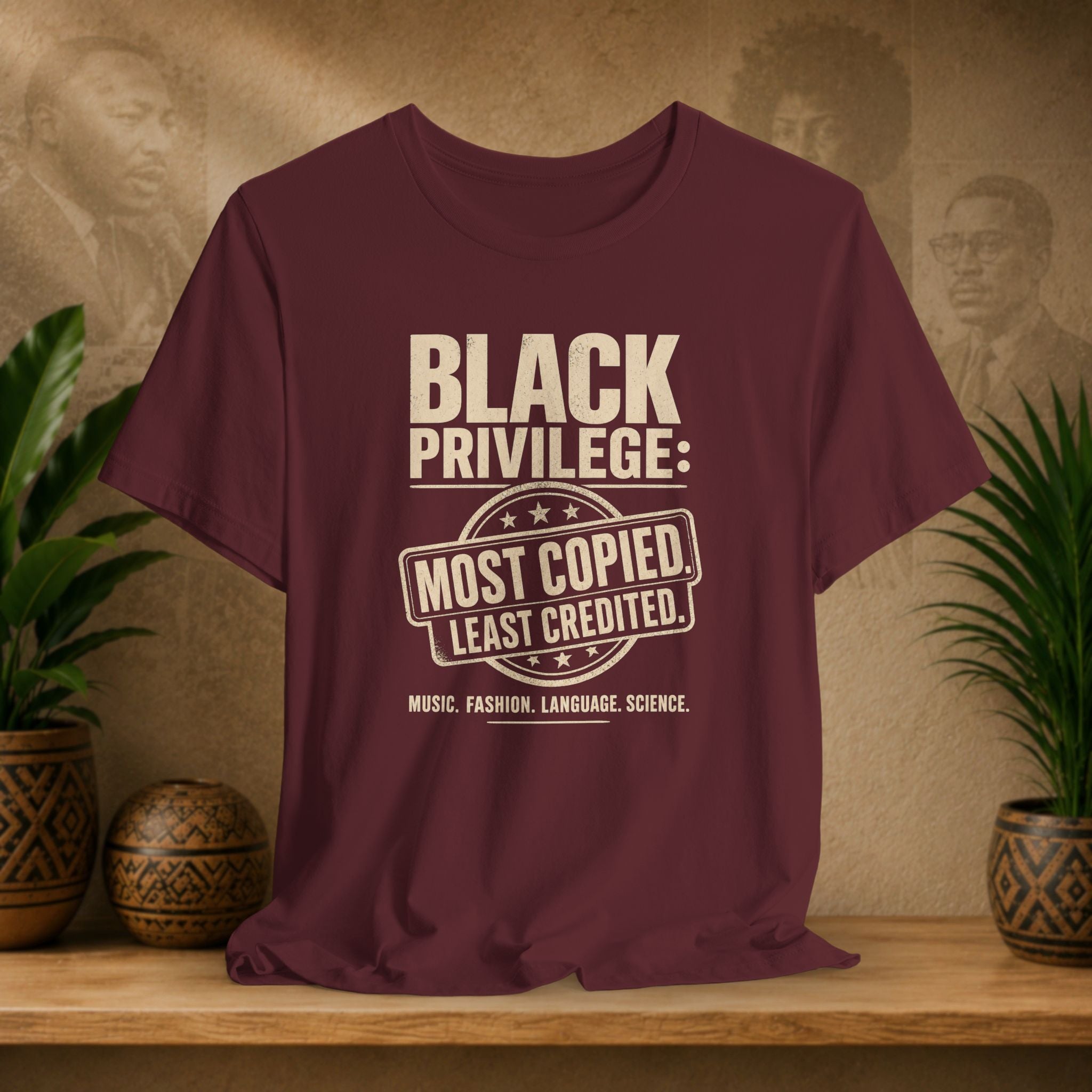 Maroon t-shirt with 'Black Privilege' text on a wooden surface with plants and a textured wall background
