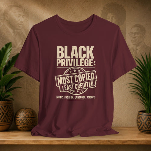 Maroon t-shirt with 'Black Privilege' text on a wooden surface with plants and a textured wall background