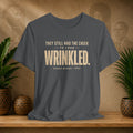 Grey t-shirt with white text on a wooden surface with plants and a textured wall background