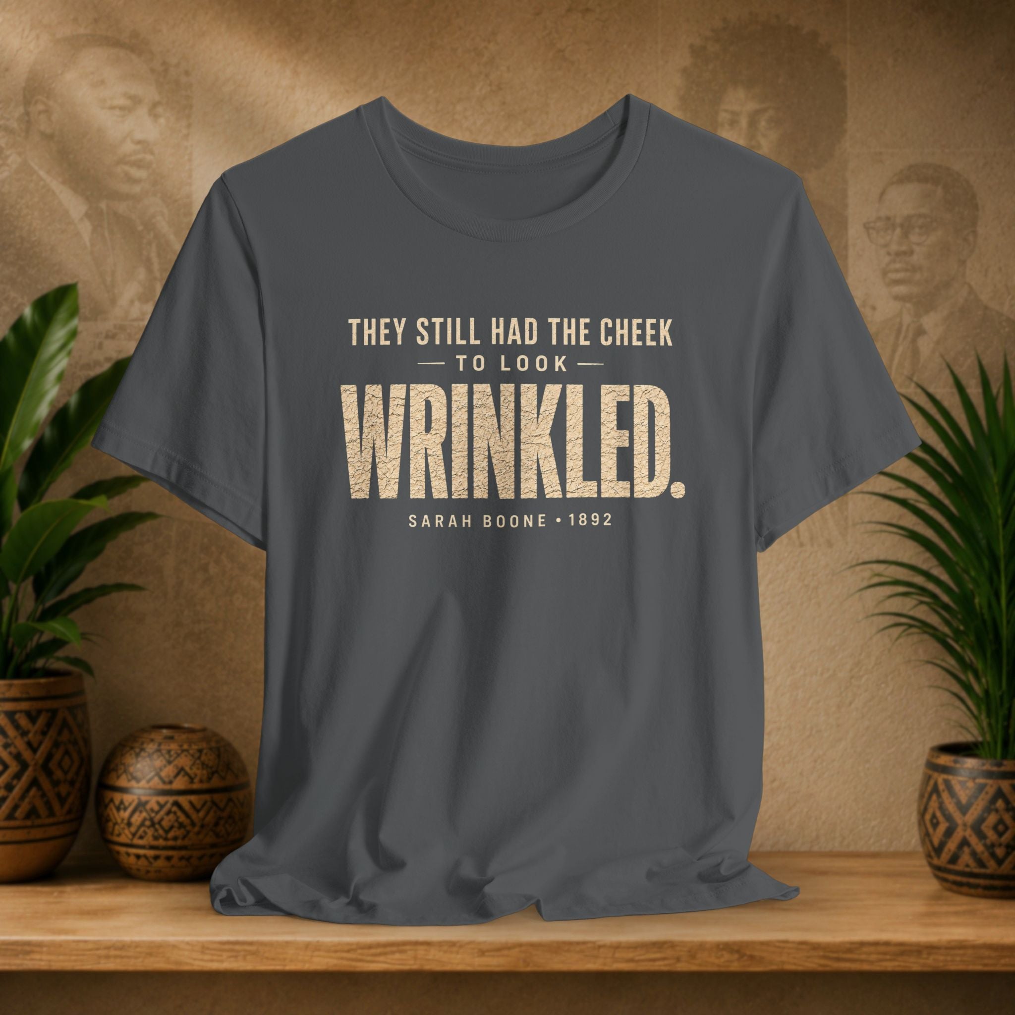 Grey t-shirt with white text on a wooden surface with plants and a textured wall background
