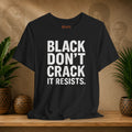 Black t-shirt with 'BLACK DON'T CRACK IT RESISTS.' text on a wooden surface with plants and a textured wall background.