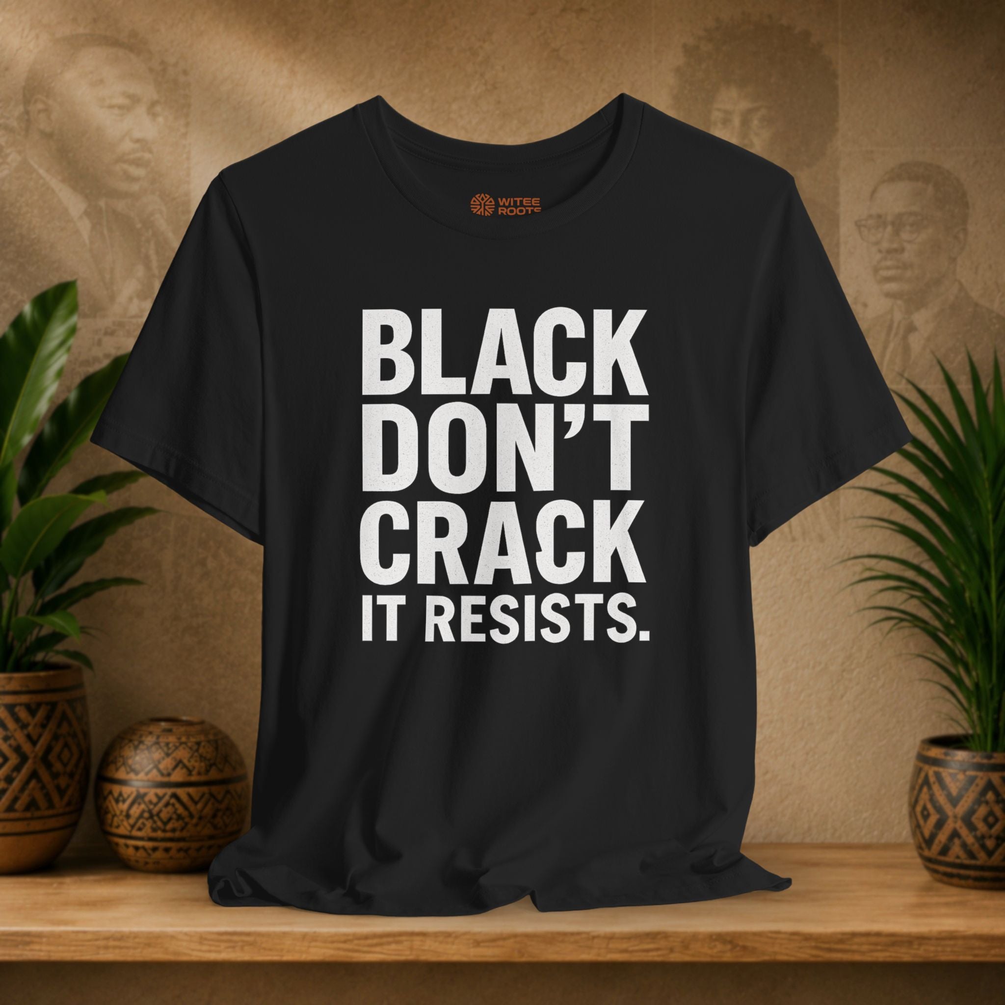 Black t-shirt with 'BLACK DON'T CRACK IT RESISTS.' text on a wooden surface with plants and a textured wall background.