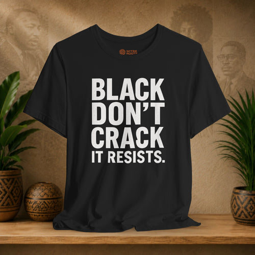 Black t-shirt with 'BLACK DON'T CRACK IT RESISTS.' text on a wooden surface with plants and a textured wall background.