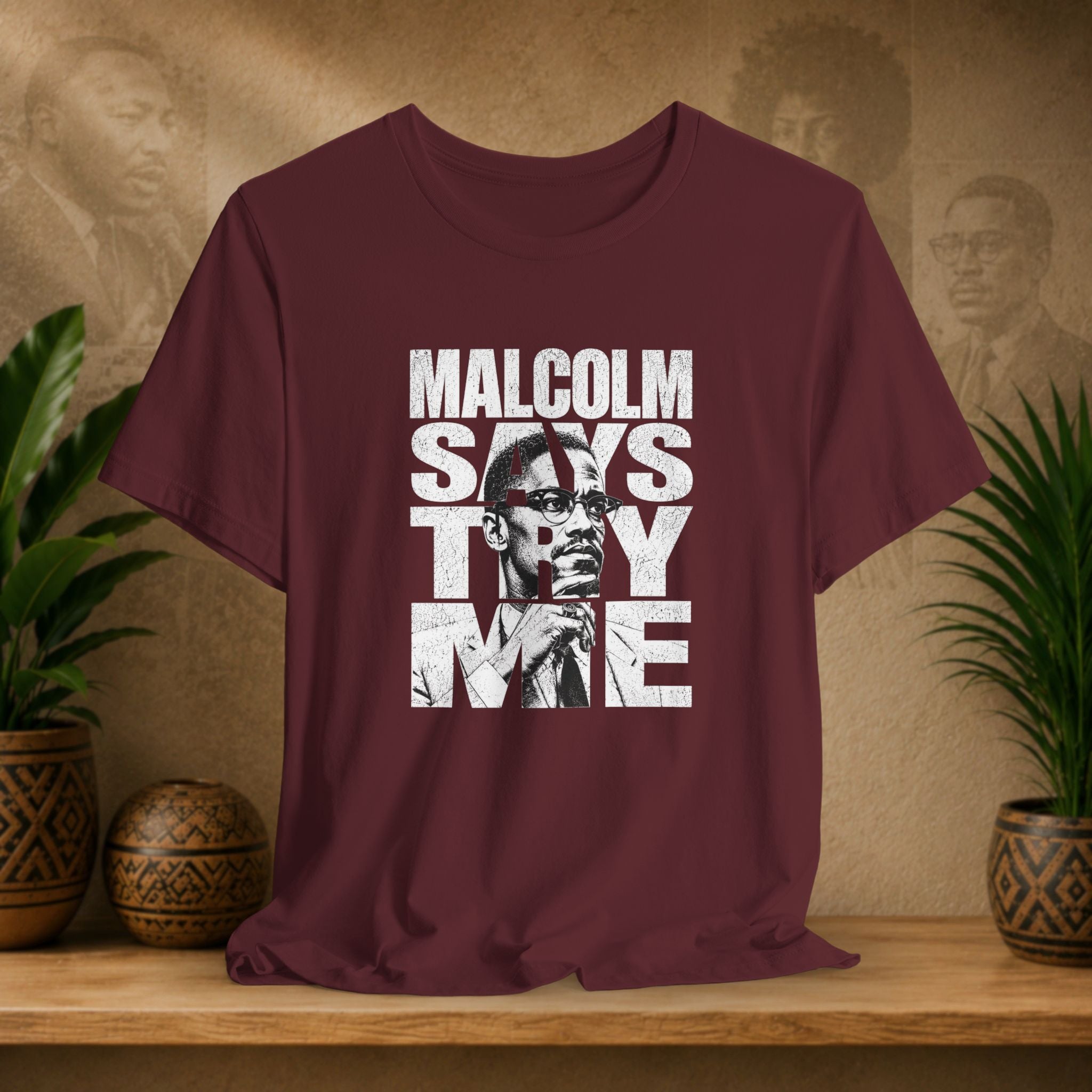 Maroon t-shirt with 'Malcolm Says Try Me' graphic on a wooden surface with plants and a textured wall in the background