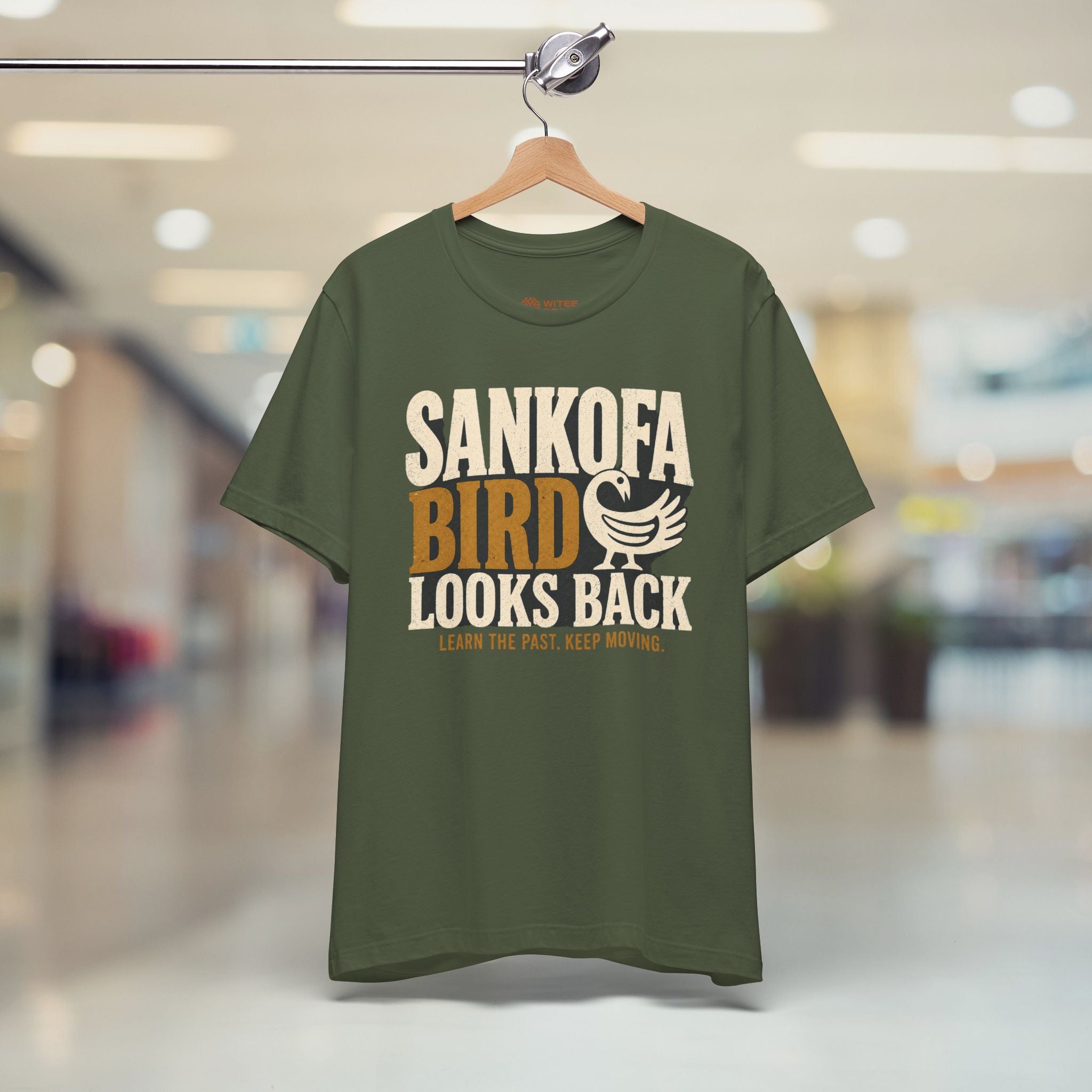 Green t-shirt with 'Sankofa Bird Looks Back' text on a hanger in a store.