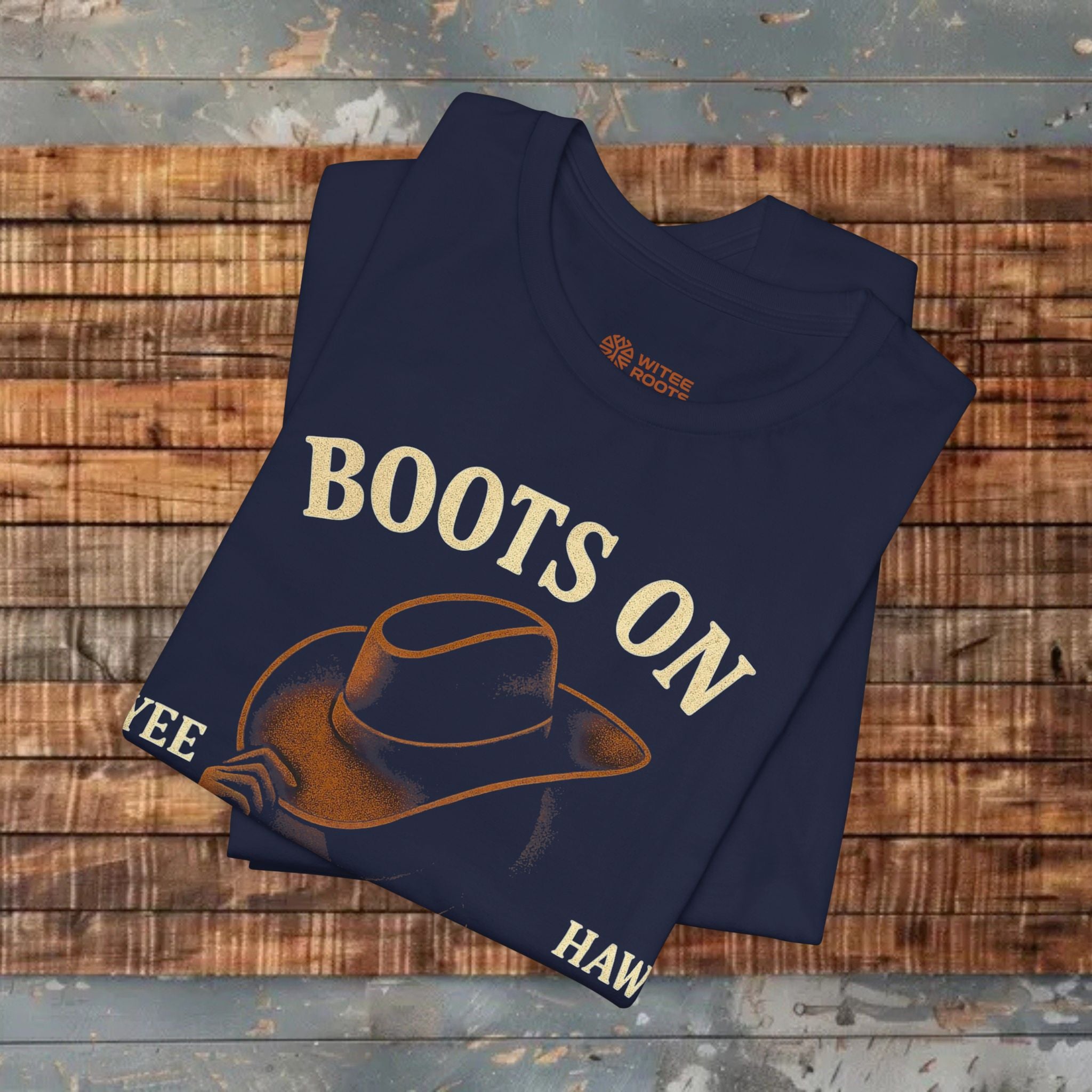 Folded navy blue t-shirt with 'BOOTS ON' text and cowboy hat graphic on a wooden surface