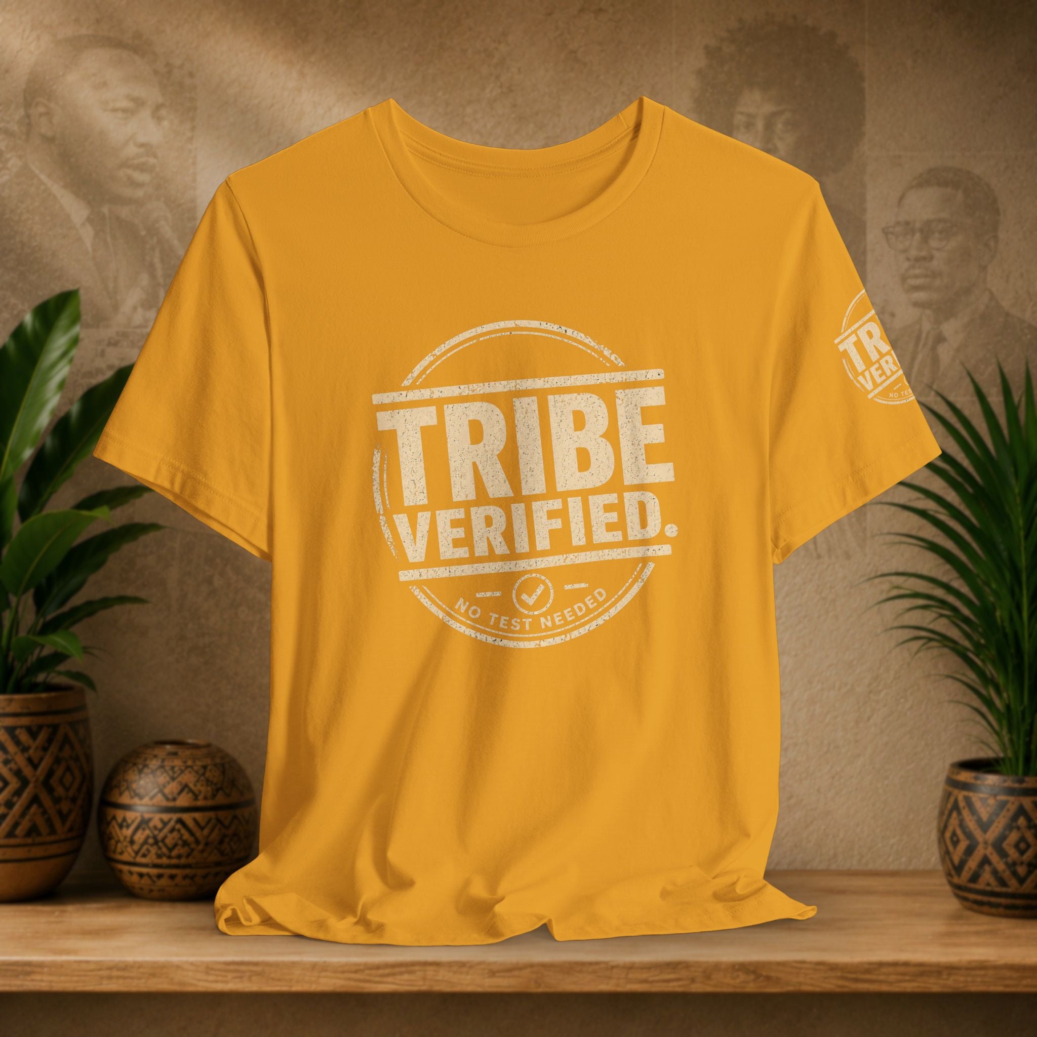 Yellow t-shirt with 'TRIBE VERIFIED' design on a wooden surface with plants and portraits in the background