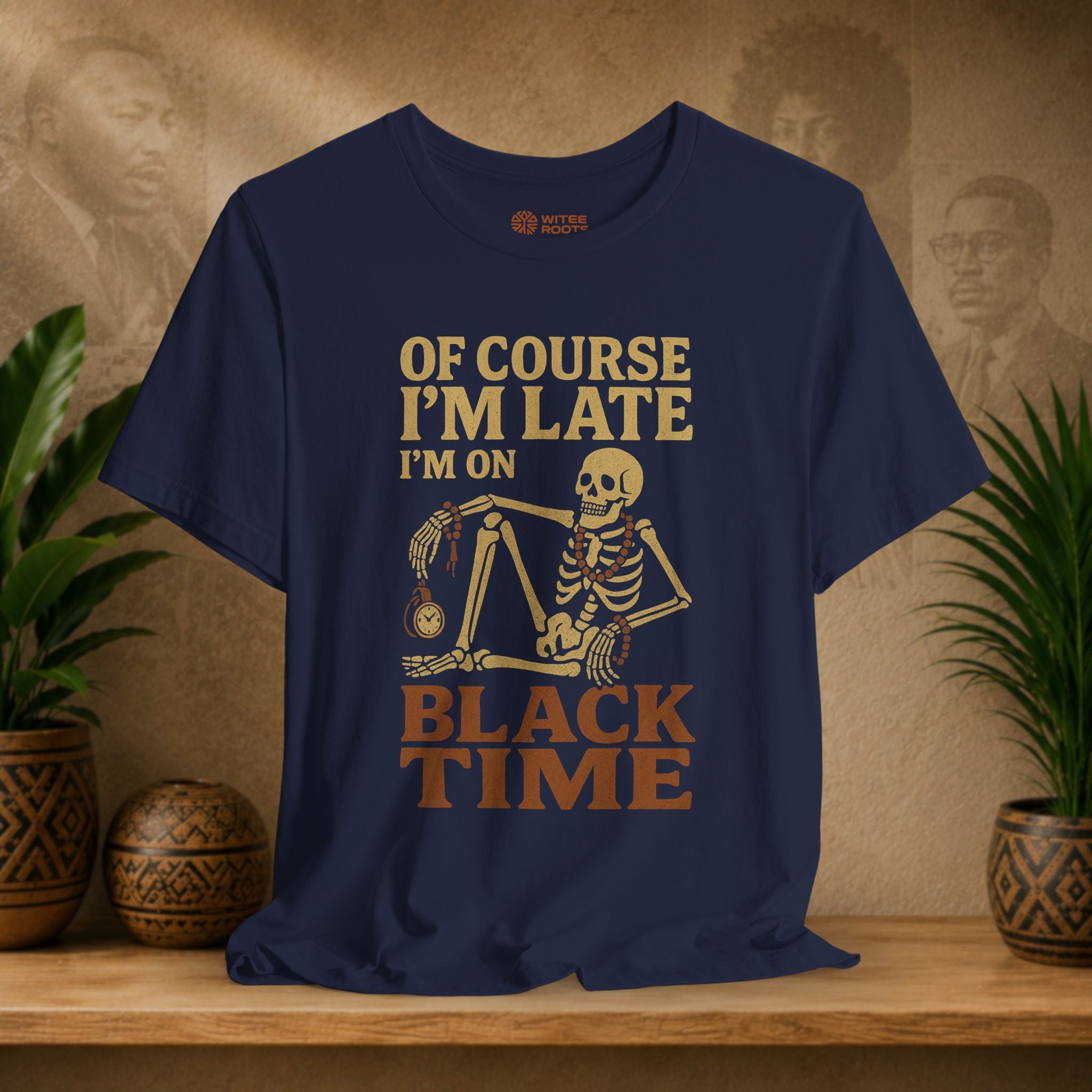A navy blue graphic t-shirt displayed on a wooden shelf. The shirt features a retro-style graphic of a skeleton holding a clock and the text 'OF COURSE I'M LATE I'M ON BLACK TIME' in tan and brown lettering.