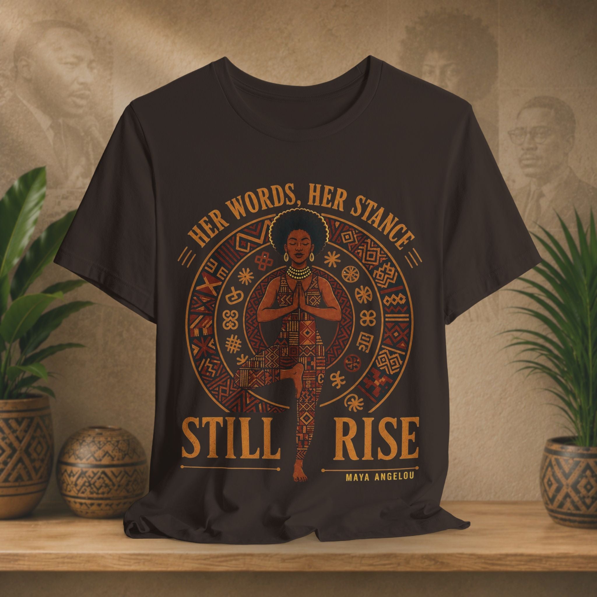 A brown charcoal gray graphic t-shirt displayed on a wooden shelf with potted plants. The shirt features a circular design with a central figure in a yoga pose, surrounded by geometric patterns and the text 'HER WORDS, HER STANCE STILL RISE' and 'MAYA ANGELUSI'.