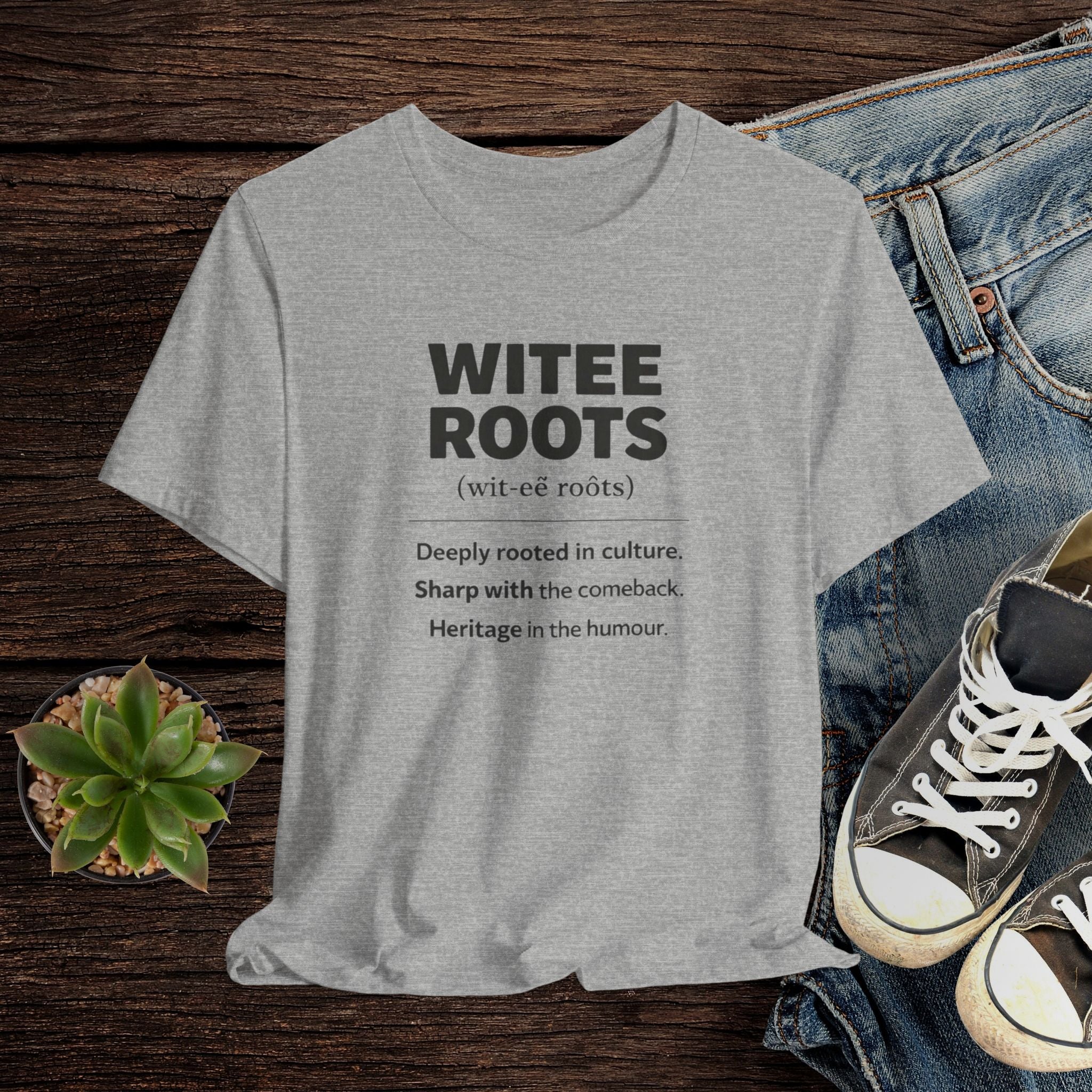 Gray t-shirt with 'WITEE ROOTS' text on a wooden surface with jeans and sneakers.