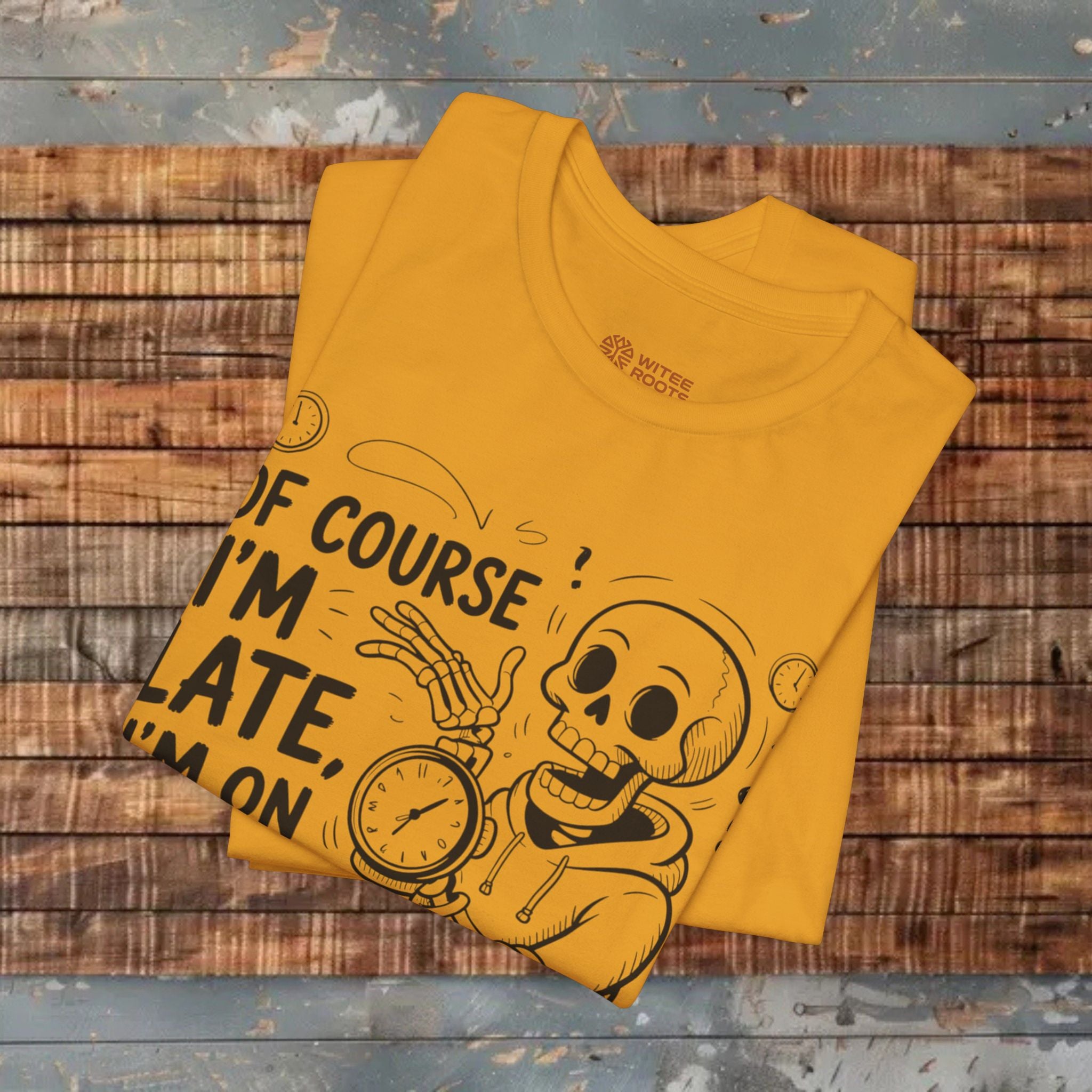 A folded mustard graphic T-shirt with a black cartoon skeleton holding a clock and the text 'OF COURSE! I'M LATE, I'M ON' is displayed on a rustic wooden plank surface.