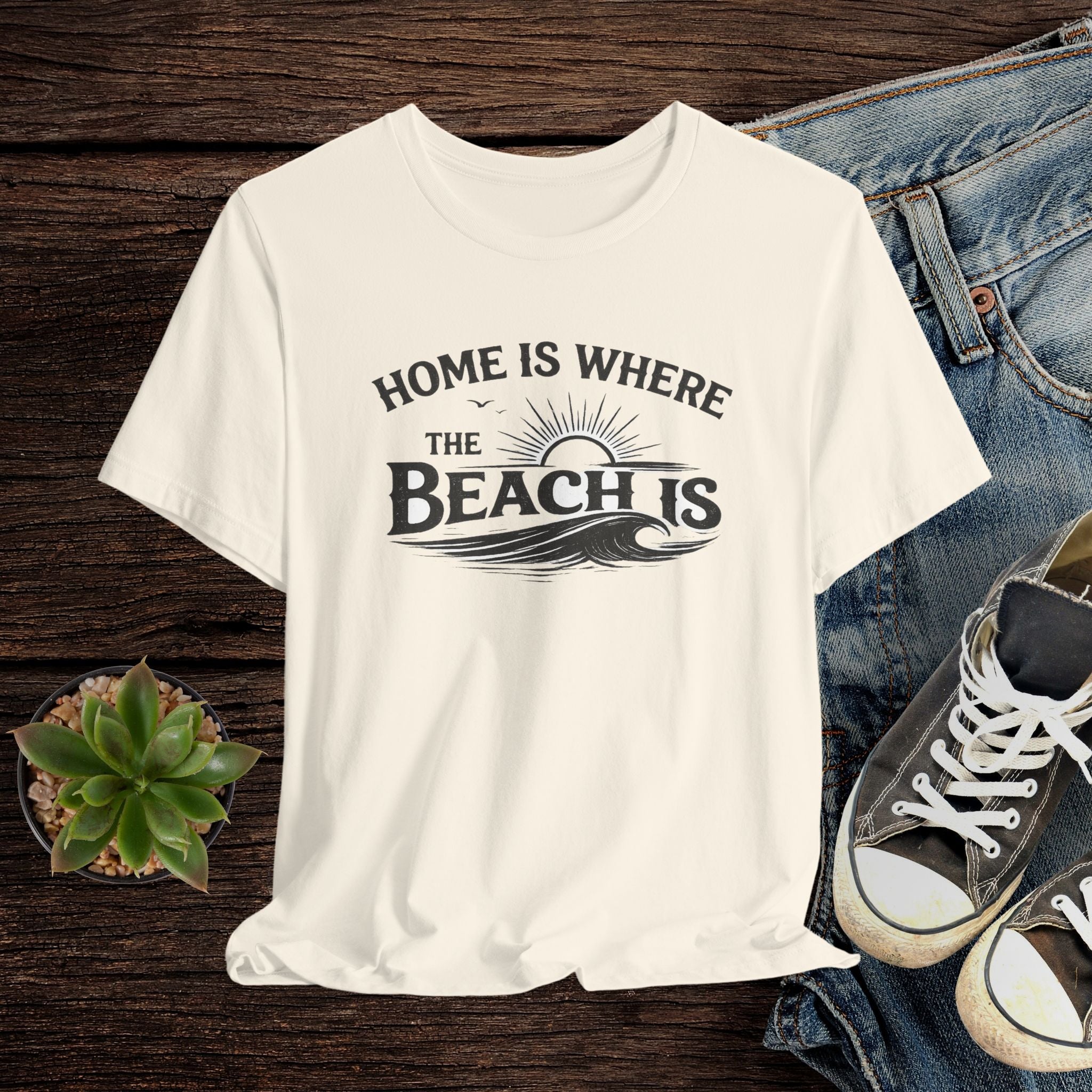 Beige t-shirt with 'Home is where the beach is' text on a wooden surface with jeans and sneakers.