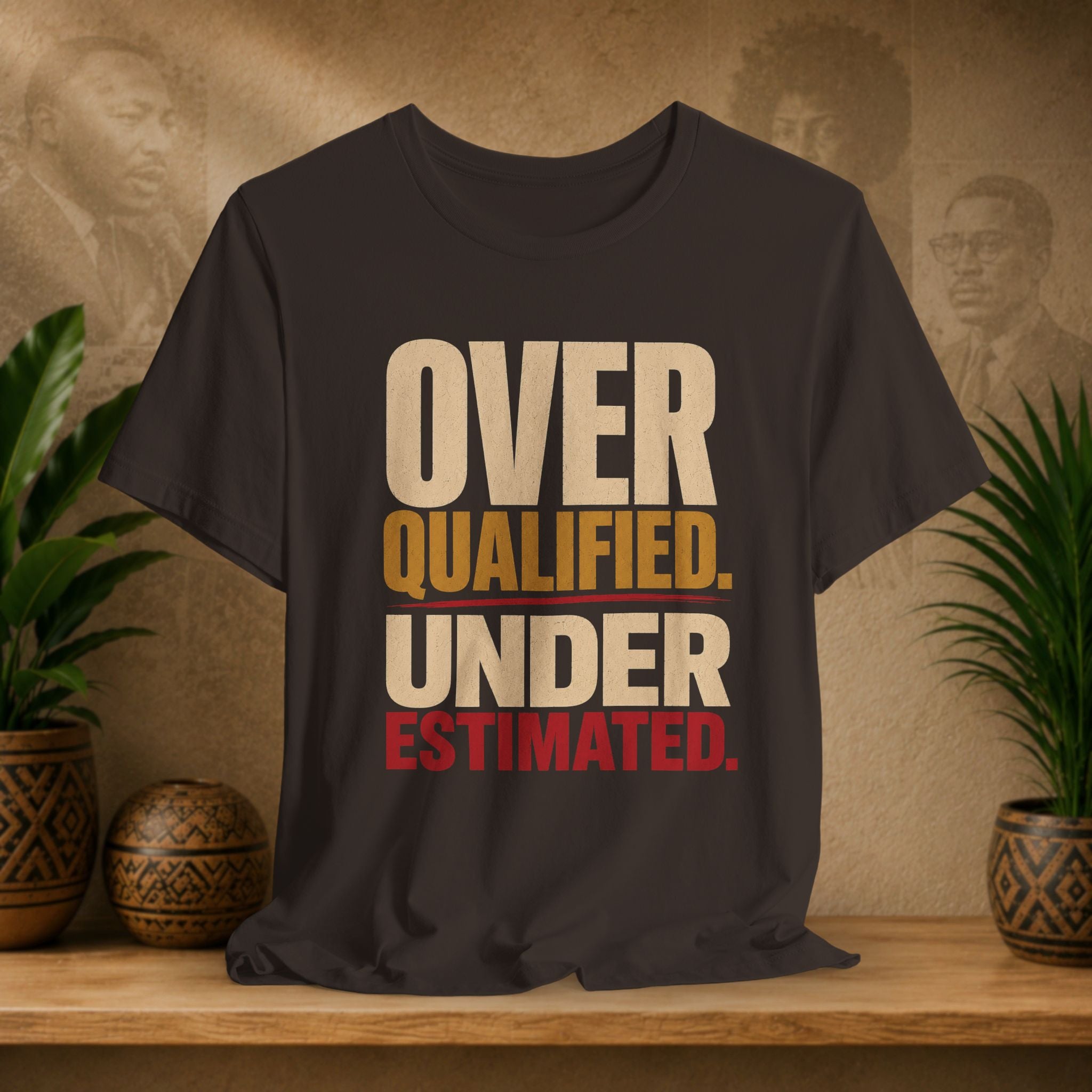 A brown graphic t-shirt displayed on a wooden shelf, flanked by potted green plants. The shirt features a bold text graphic reading 'OVER QUALIFIED.