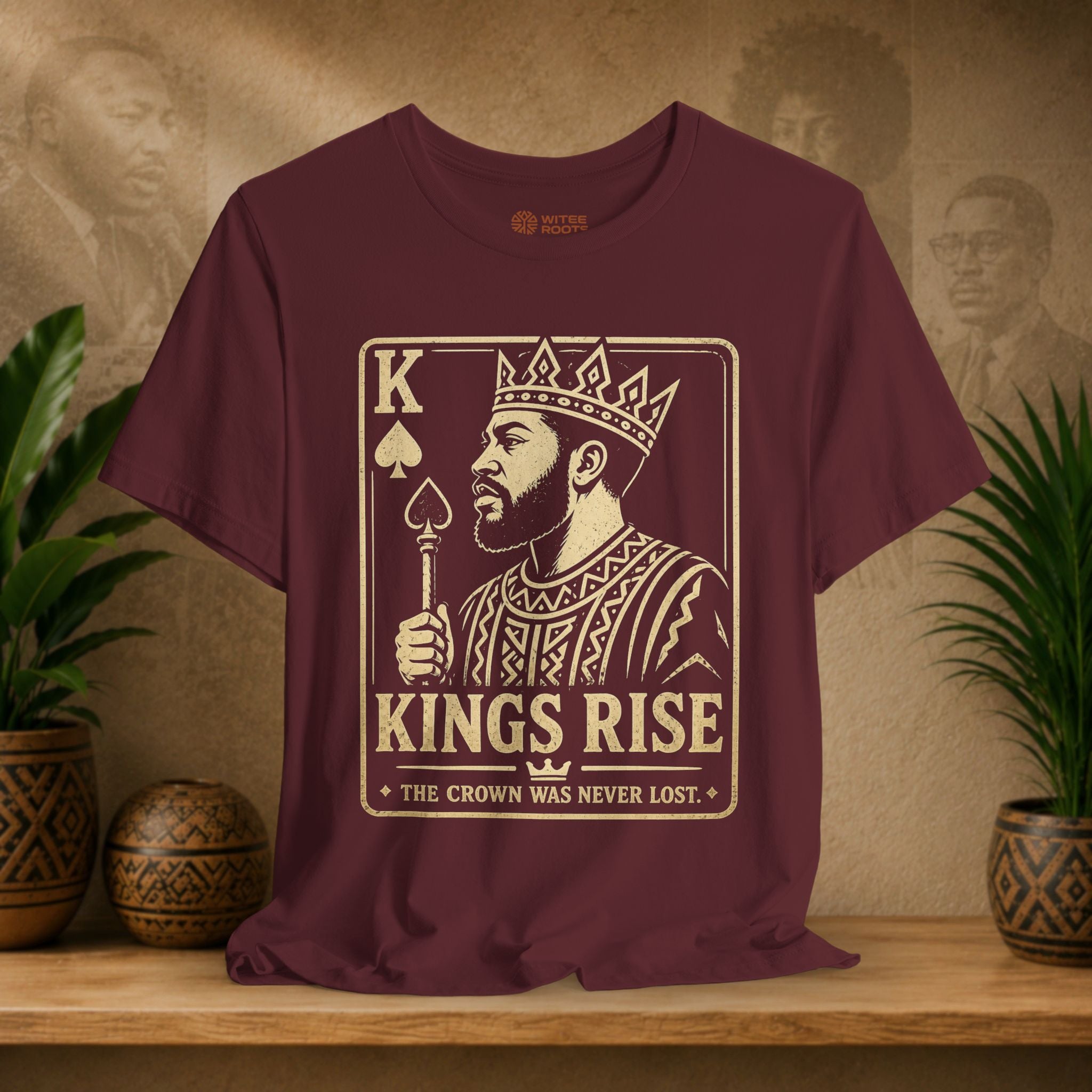 Maroon t-shirt with a graphic design of a king holding a crown and text on a wooden surface with plants and a decorative item in the background.