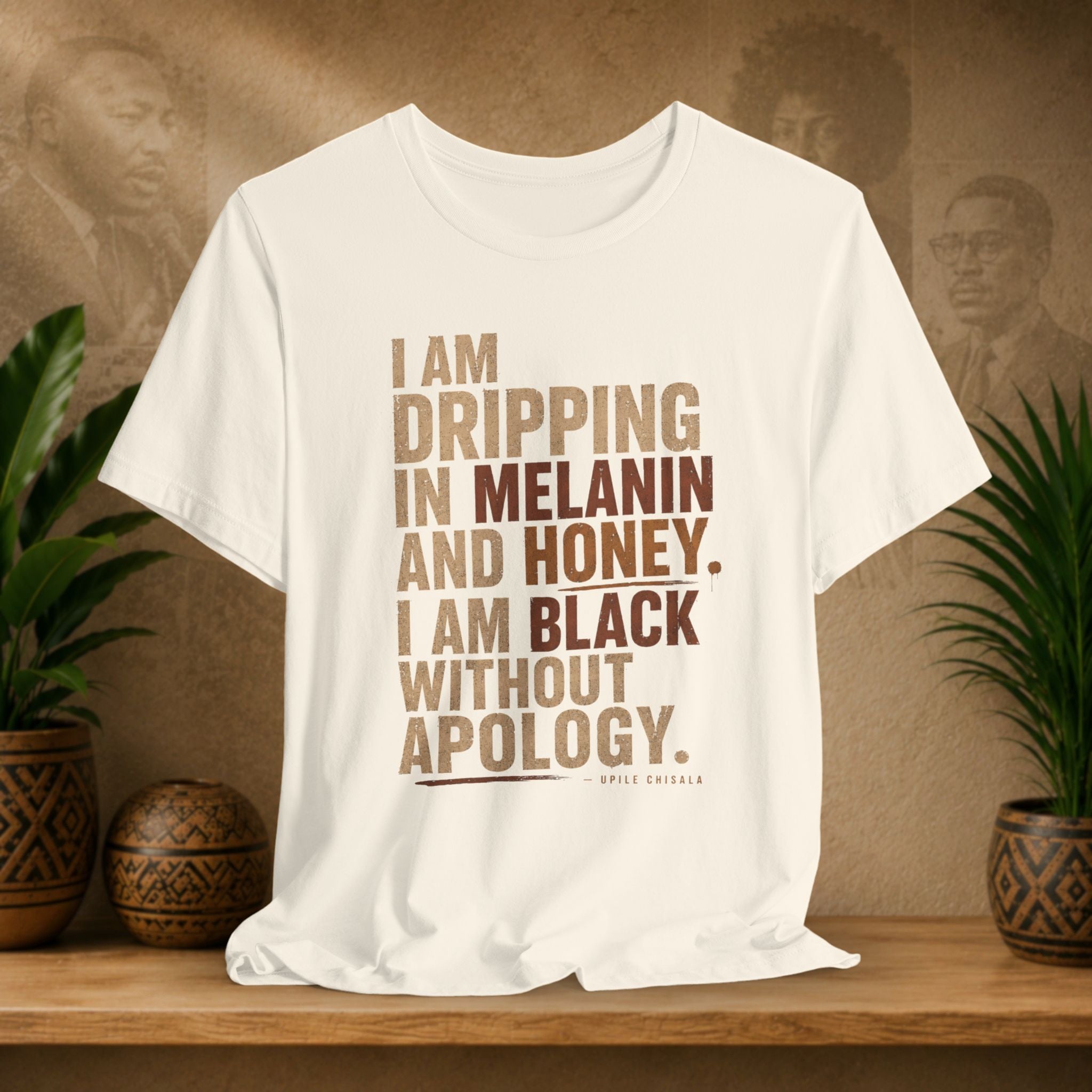 A off-white short-sleeve graphic T-shirt displayed on a wooden surface against a warm brown backdrop with decorative plants. The shirt features stacked text reading 'I AM DRIPPING IN MELANIN AND HONEY.