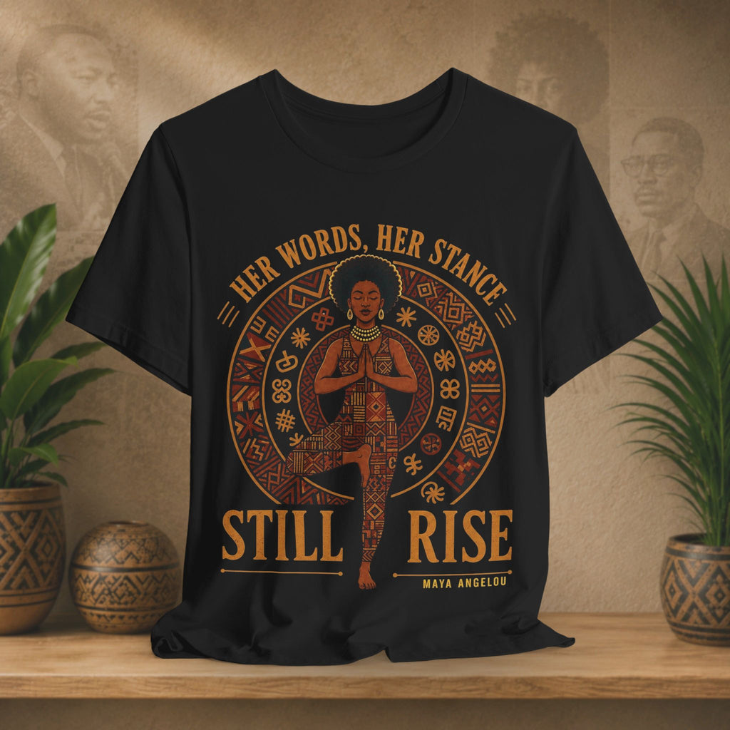 A black short-sleeve graphic T-shirt displayed on a wooden shelf, featuring a circular Native American-inspired design with a central figure and the text 'HER WORDS, HER STANCE STILL RISE' and 'MAYA ANGELOS'.