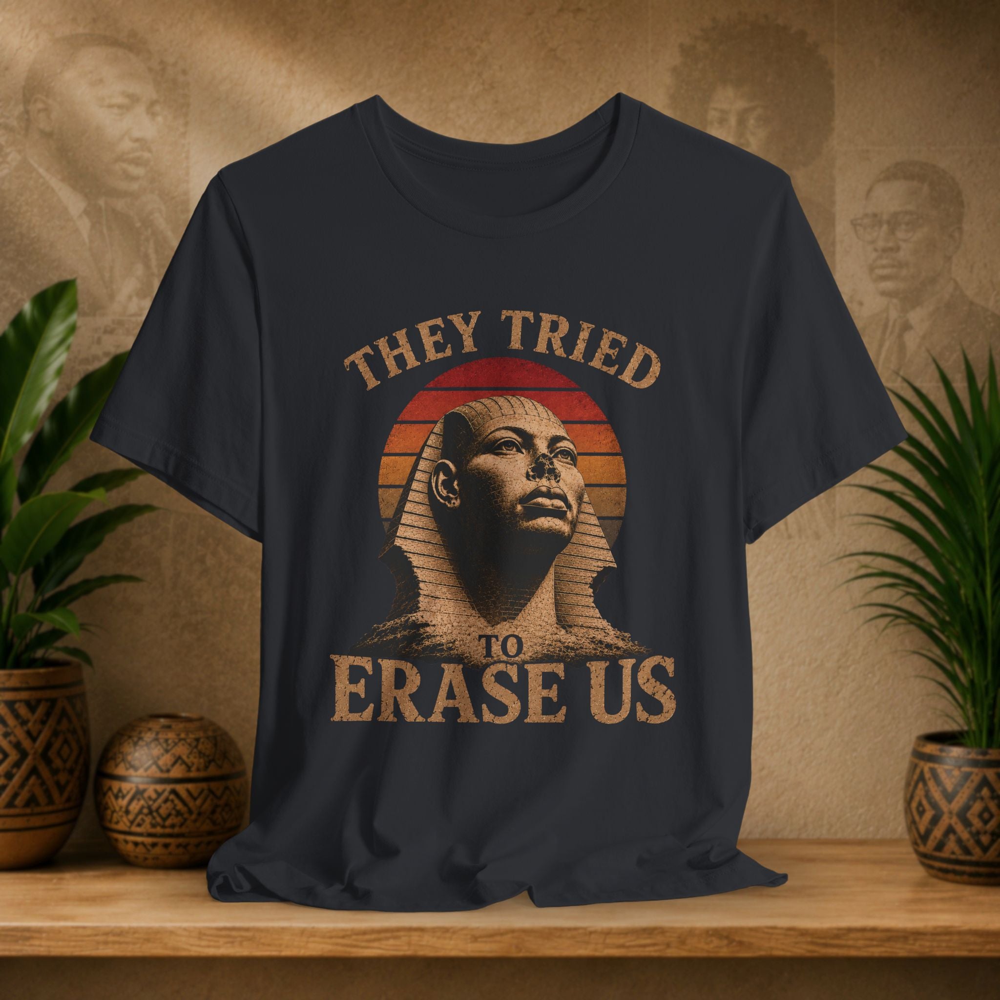 Black t-shirt with a graphic of a sphinx and text 'They Tried To Erase Us' on a wooden surface with plants and decorative items.