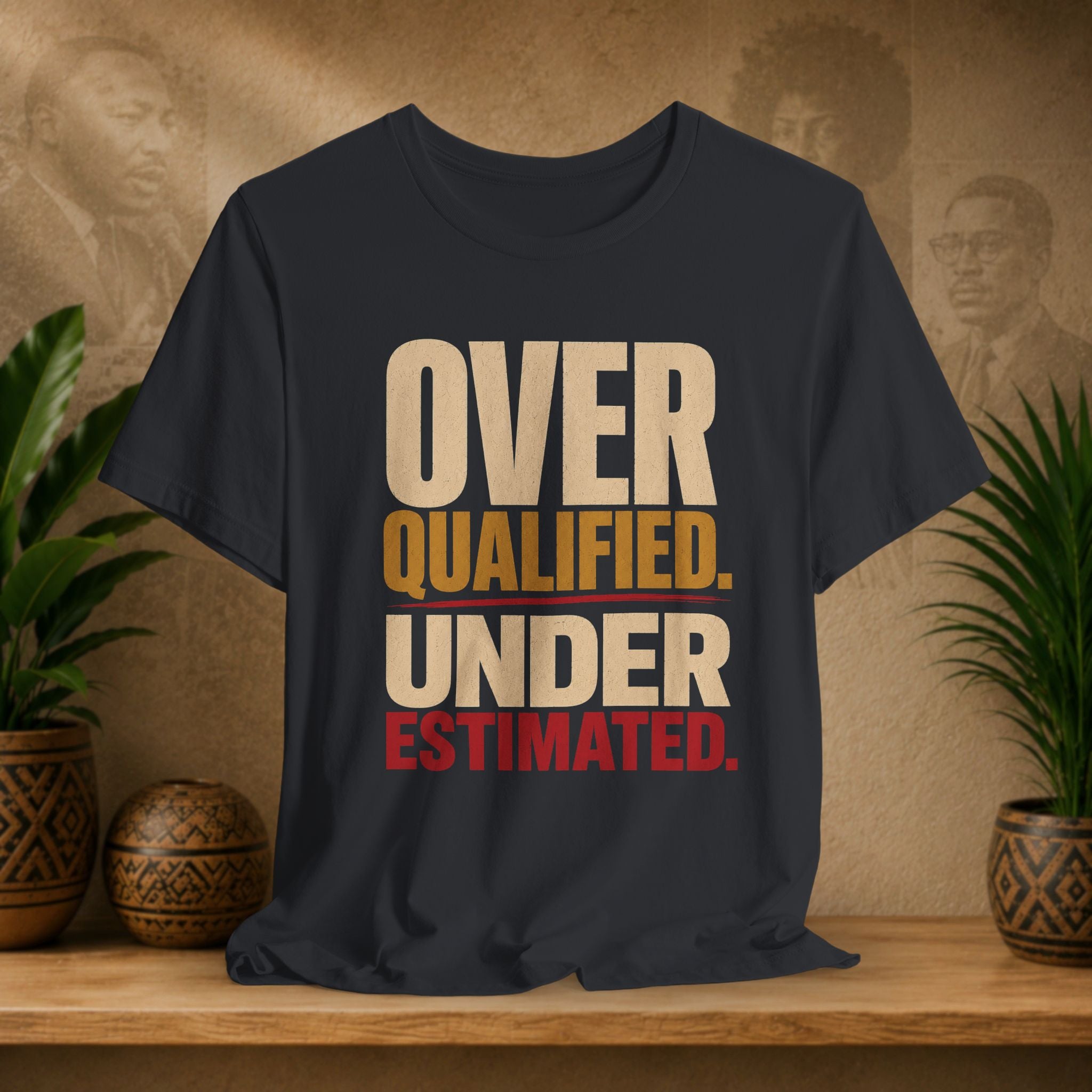 A black graphic t-shirt displayed on a wooden shelf, flanked by potted green plants. The shirt features a bold text graphic reading 'OVER QUALIFIED.