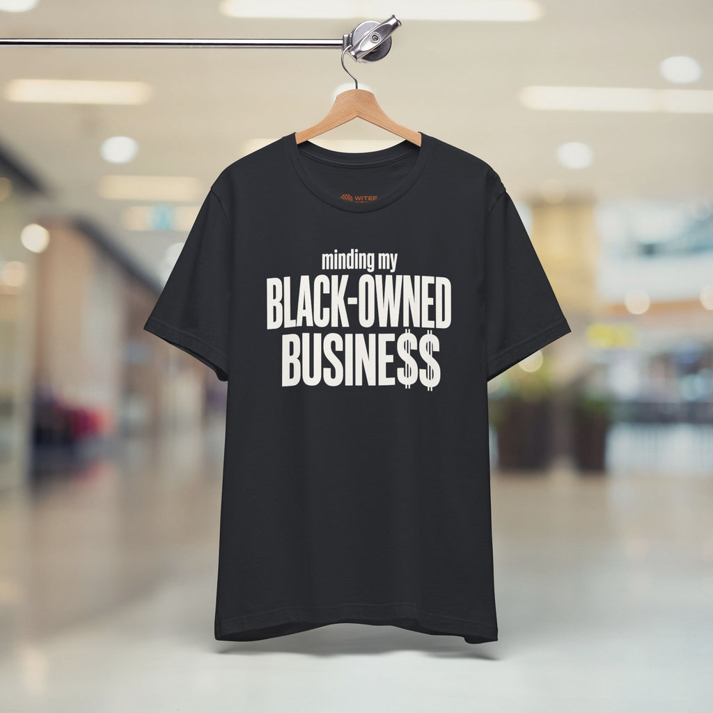 A black short-sleeve t-shirt hanging on a wooden hanger from a clothing rack, featuring the text 'minding my BLACK-OWNED BUSINESS$$' in white. The background is a blurred indoor shopping mall corridor.