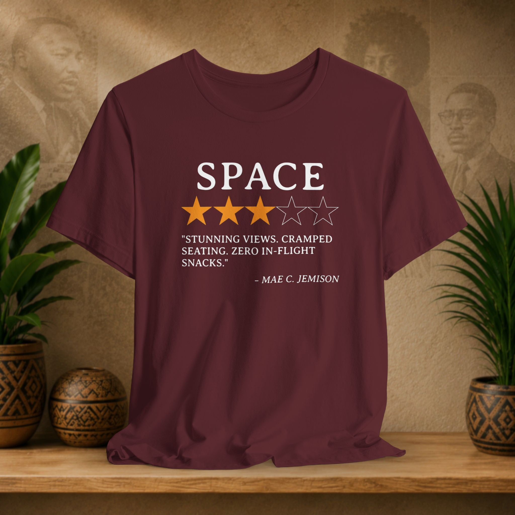 Maroon t-shirt with space-themed text and rating on a wooden surface with plants and wall art in the background.