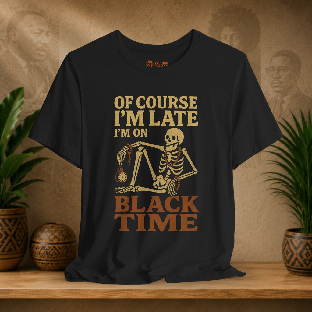 A black short-sleeve graphic T-shirt displayed on a wooden shelf, featuring a tan and brown skeleton sitting on a globe with the text 'OF COURSE I'M LATE I'M ON BLACK TIME'. The background is a warm brown wall with sepia portraits, flanked by potted green plants.