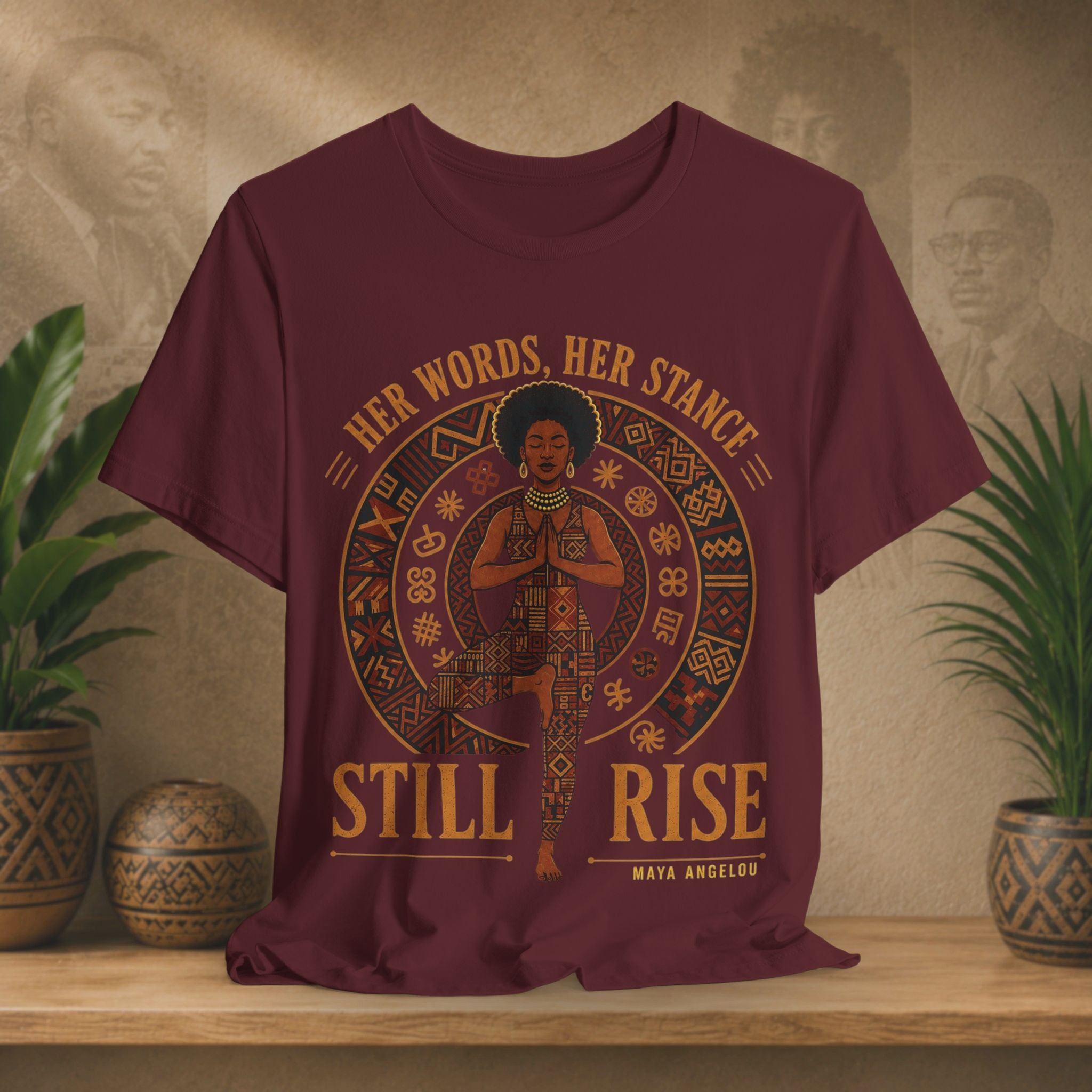 A maroon short-sleeve graphic T-shirt displayed on a wooden shelf, flanked by potted plants. The shirt features a circular graphic of a woman in traditional attire with the text 'HER WORDS, HER STANCE STILL RISE' and 'MAYA ANGELUS' printed below.