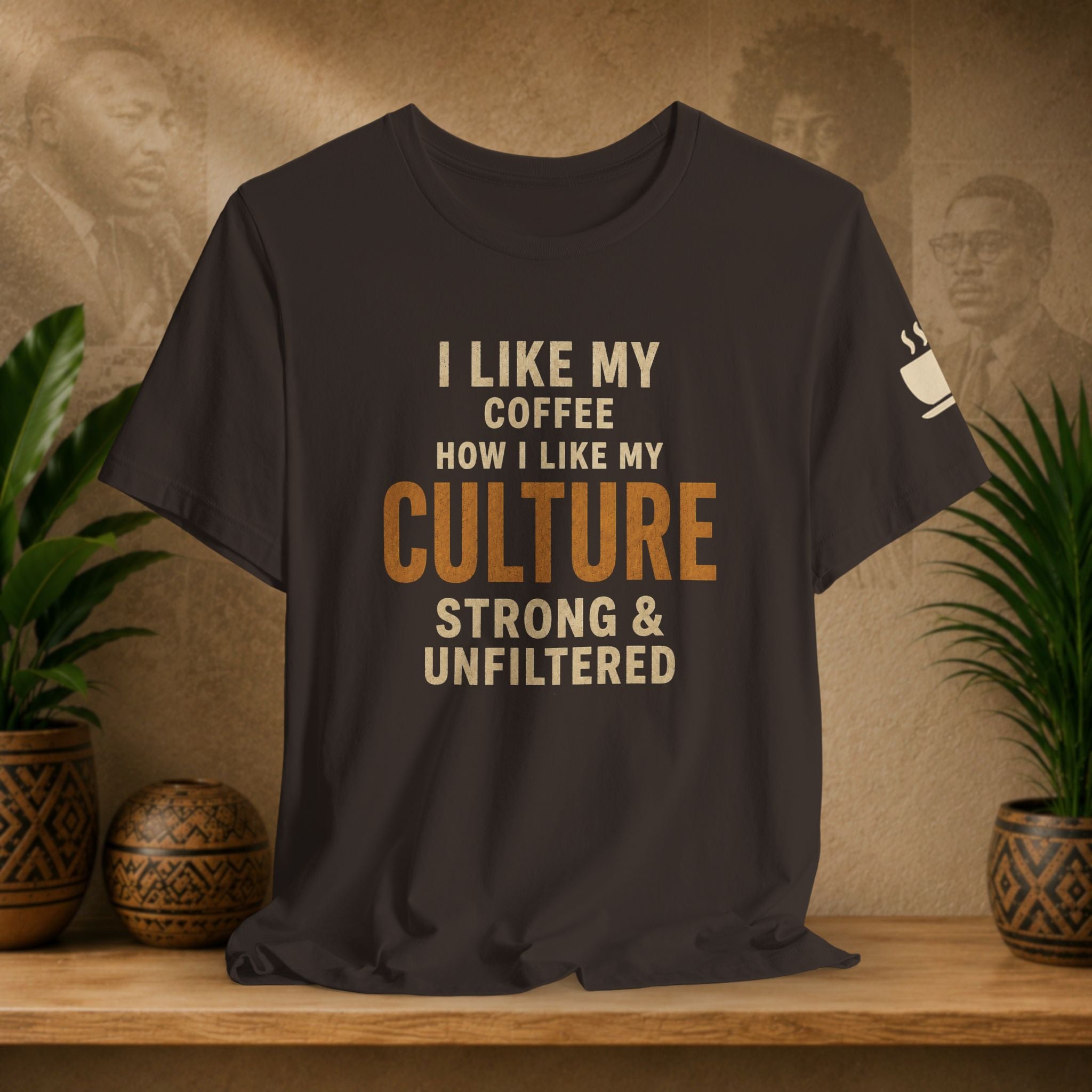 A brown graphic t-shirt displayed on a wooden shelf, flanked by potted green plants. The shirt features the text 'I LIKE MY COFFEE HOW I LIKE MY CULTURE STRONG & UNFILTERED' in cream and orange lettering, with a small white coffee cup graphic on the left sleeve.