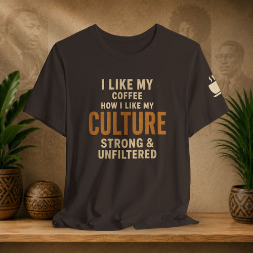 A brown graphic t-shirt displayed on a wooden shelf, flanked by potted green plants. The shirt features the text 'I LIKE MY COFFEE HOW I LIKE MY CULTURE STRONG & UNFILTERED' in cream and orange lettering, with a small white coffee cup graphic on the left sleeve.