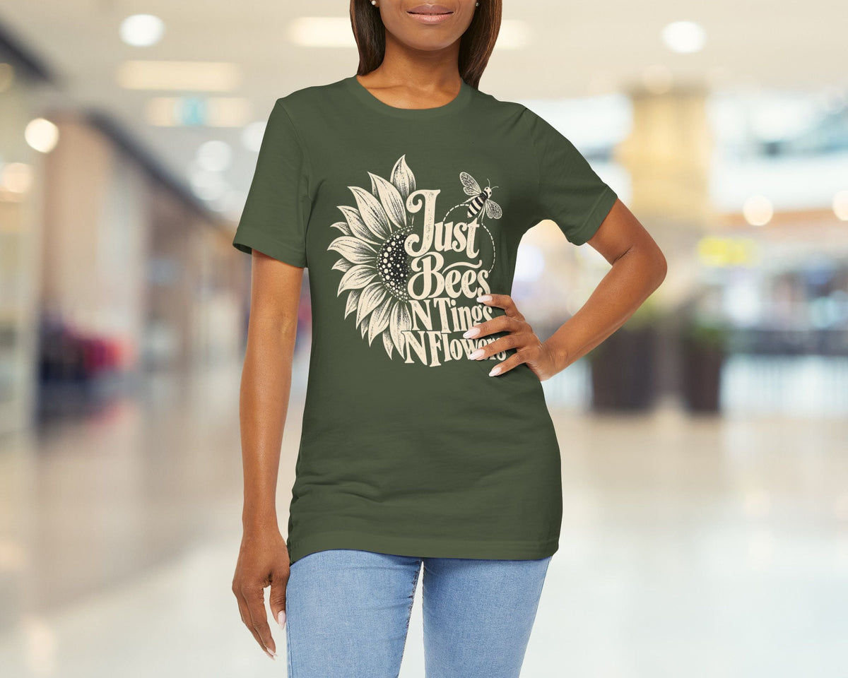 A woman modeling a dark olive green t-shirt with a white graphic that reads 'Just Bees Never Flowers' around a sunflower and bee design, set against a blurred indoor hallway background.