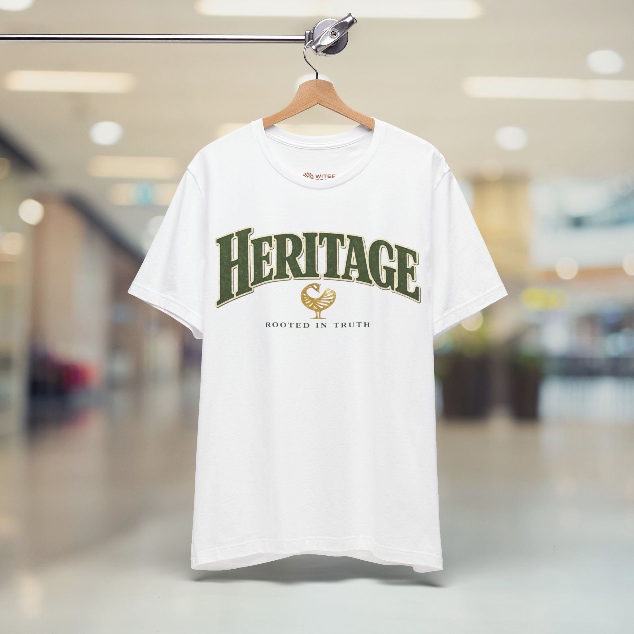 White t-shirt with 'Heritage' text on a hanger in a blurred indoor setting