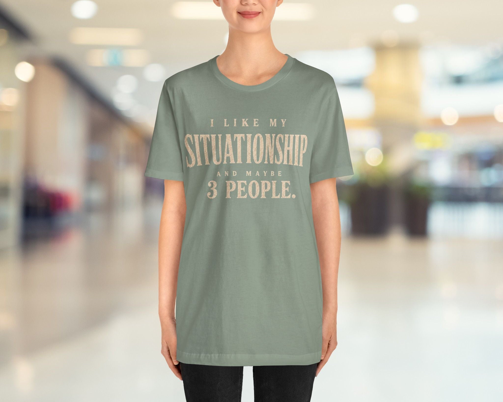 A woman modeling a sage green t-shirt with a beige graphic print that reads 'I LIKE MY SITUATIONSHIP FOR MAYBE 3 PEOPLE.' against a blurred indoor hallway background.