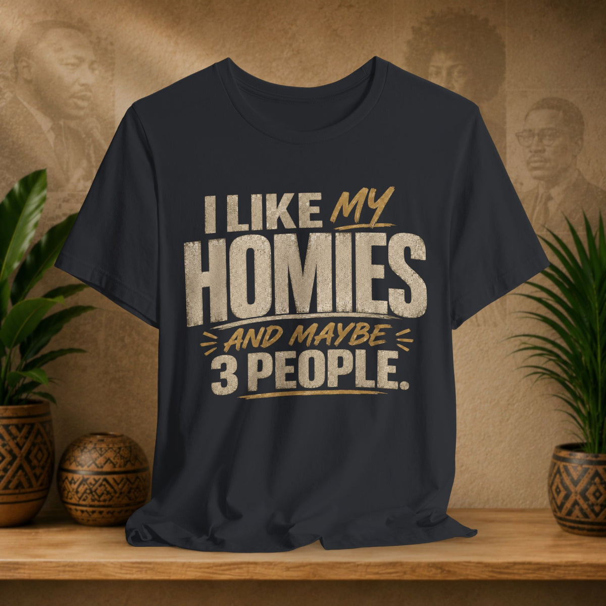 A black graphic t-shirt displayed on a wooden shelf. The shirt features a humorous slogan printed in white and tan lettering: 'I LIKE MY HOMIES and maybe 3 PEOPLE.