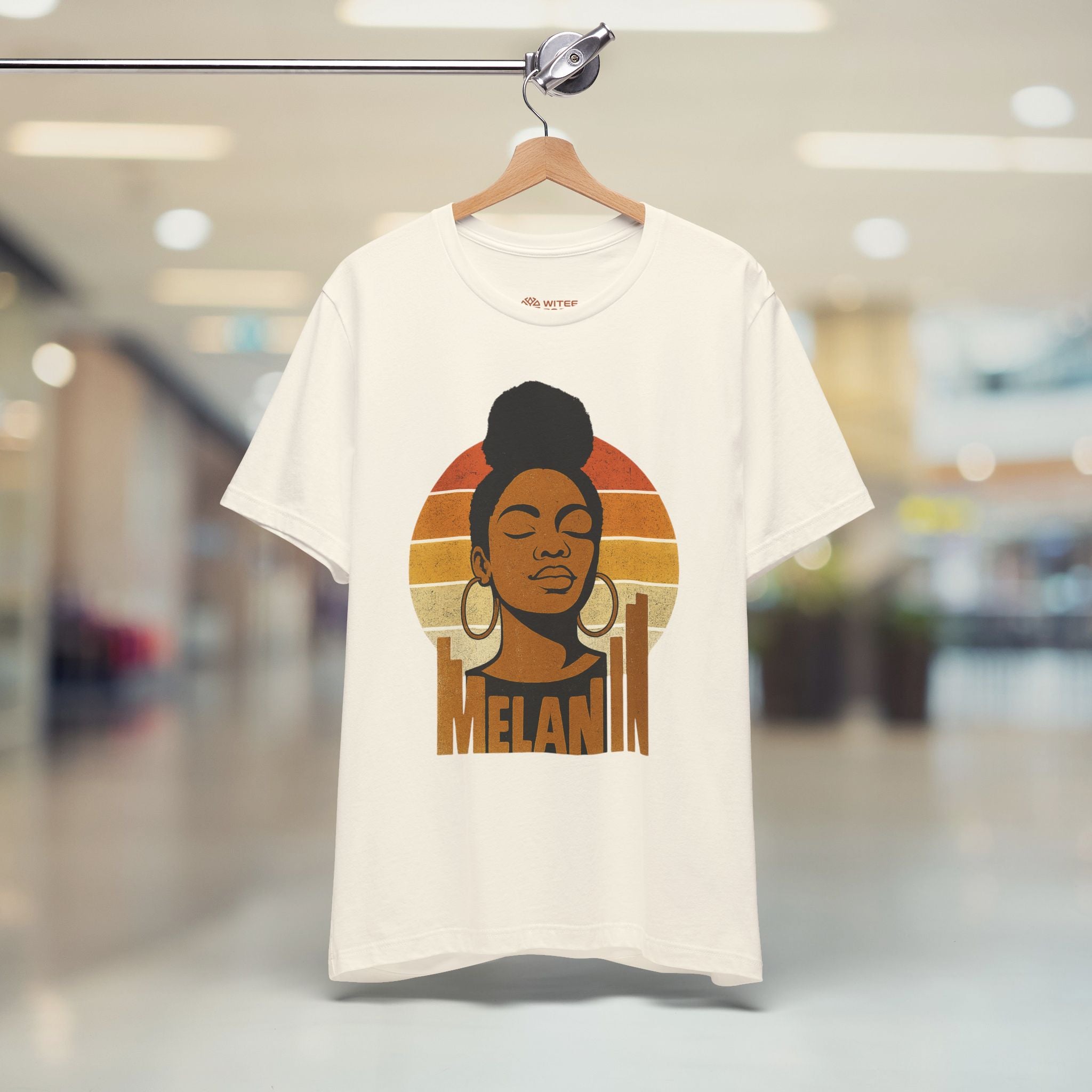 Cream t-shirt with a graphic design of a woman's face and 'Melanin' text, hanging in a store.