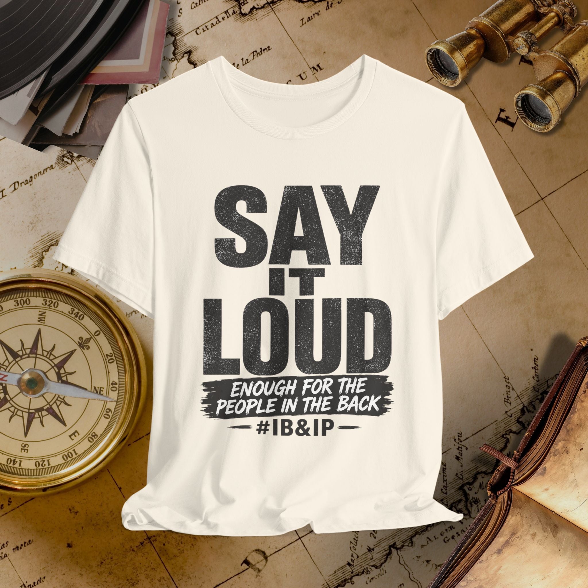 Beige t-shirt with text 'SAY IT LOUD ENOUGH FOR THE PEOPLE IN THE BACK #IB&IP' on a vintage map background.