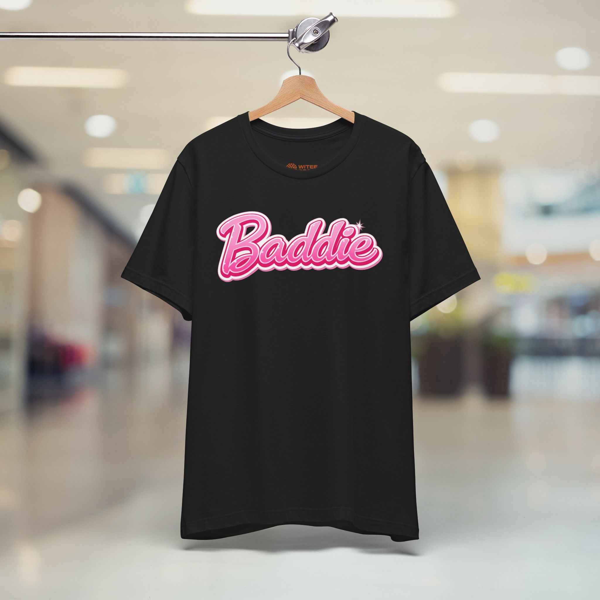 A black short-sleeve t-shirt hanging on a wooden hanger from a clothing rack, featuring a pink 'Baddie' graphic across the chest, set against a blurred indoor hallway background.