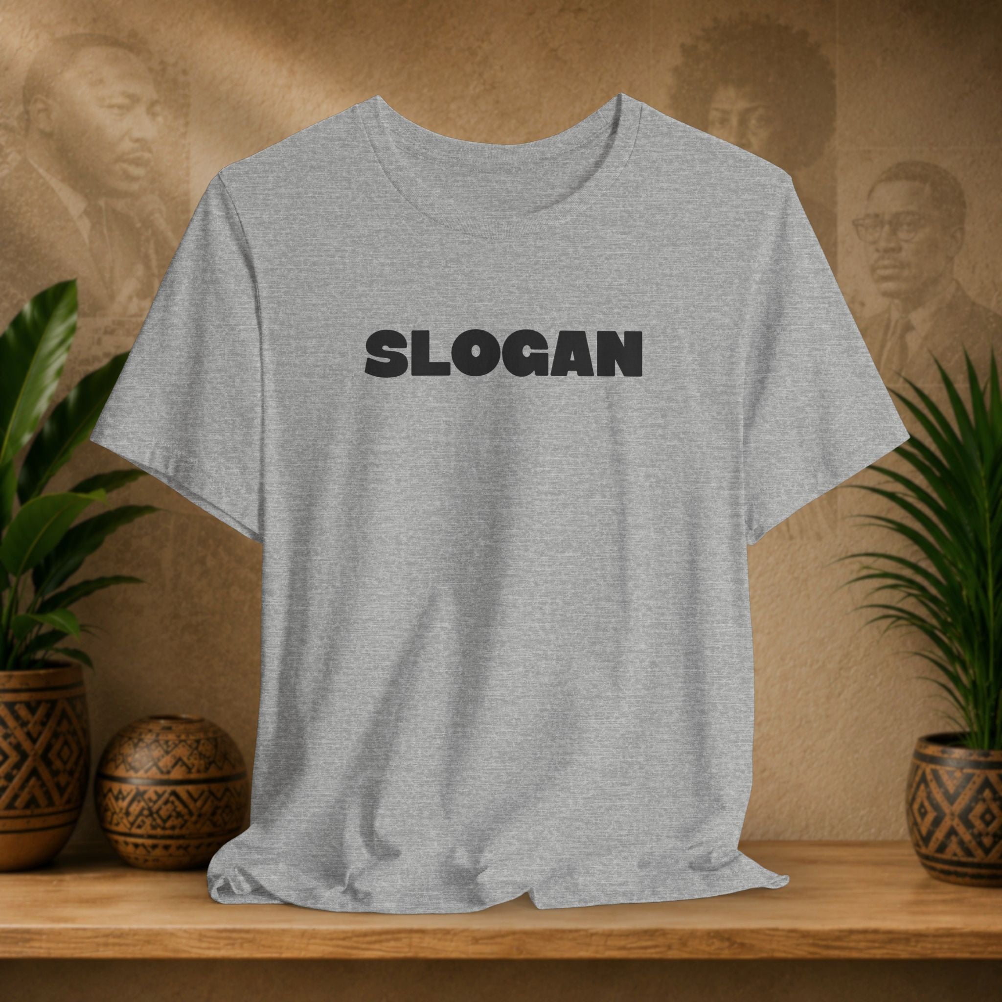 A light heather gray short-sleeve t-shirt with the word 'SLOGAN' printed in bold black capital letters across the chest, displayed on a wooden shelf with potted plants and a textured background.